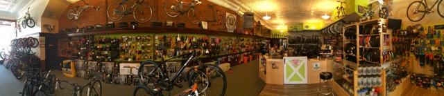 Panoramic view of a bicycle shop featuring a variety of bikes displayed on walls and the floor, colorful accessories on shelves, and a brightly lit sales counter. The interior has a warm ambiance with brick walls and green accents.