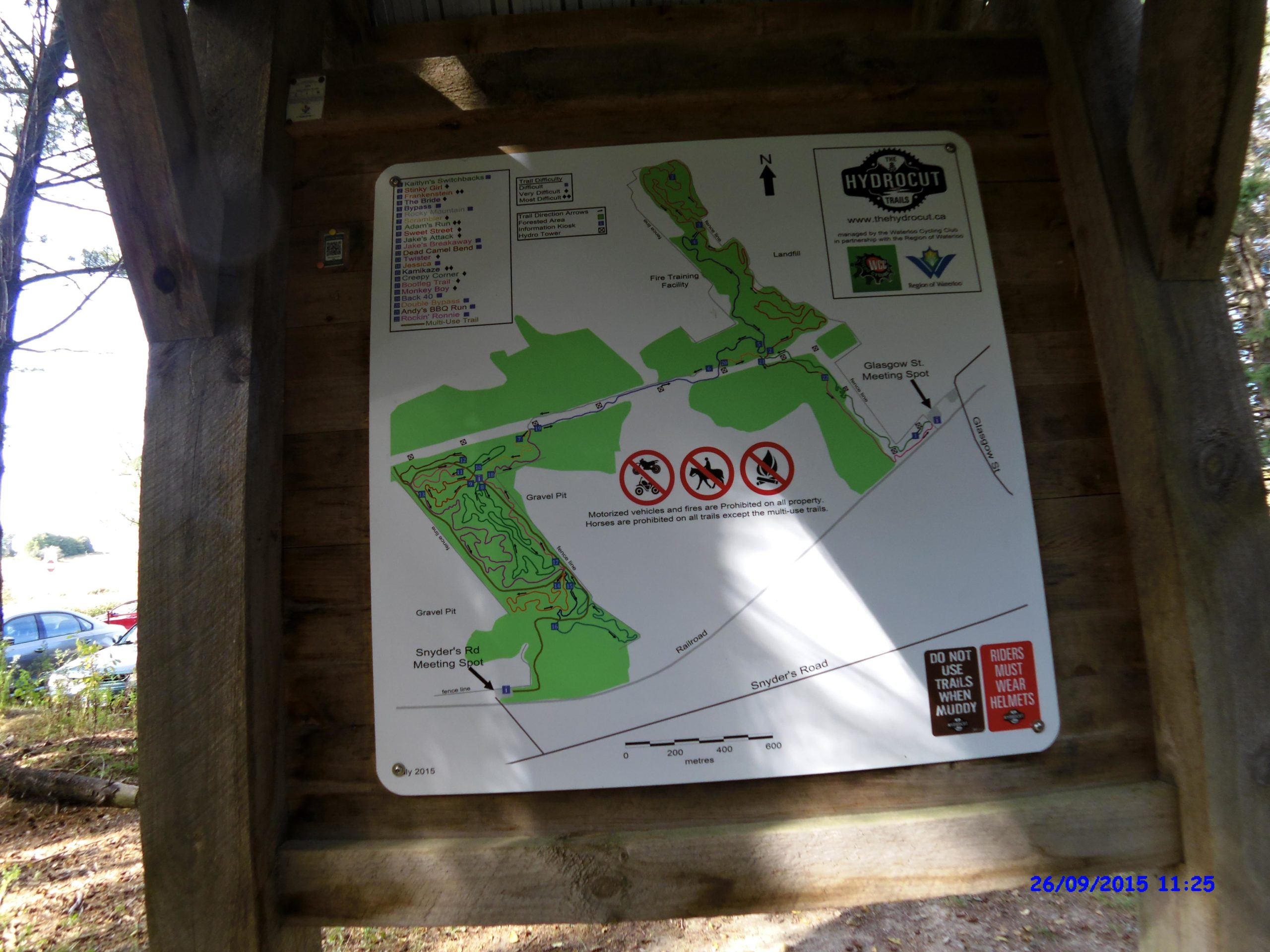 Map of the Hydrocut trails, displaying various trails, meeting points, and regulations. The map includes marked areas like "Gravel Pit" and "Fire Training Facility," along with symbols indicating trails for different uses and areas where motorized vehicles and horses are prohibited. Warning notes emphasize not to use trails when muddy and the requirement for riders to wear helmets. The image is captured from a wooden shelter with a partially visible wooded area in the background. Hydrocut mountain bike trail.