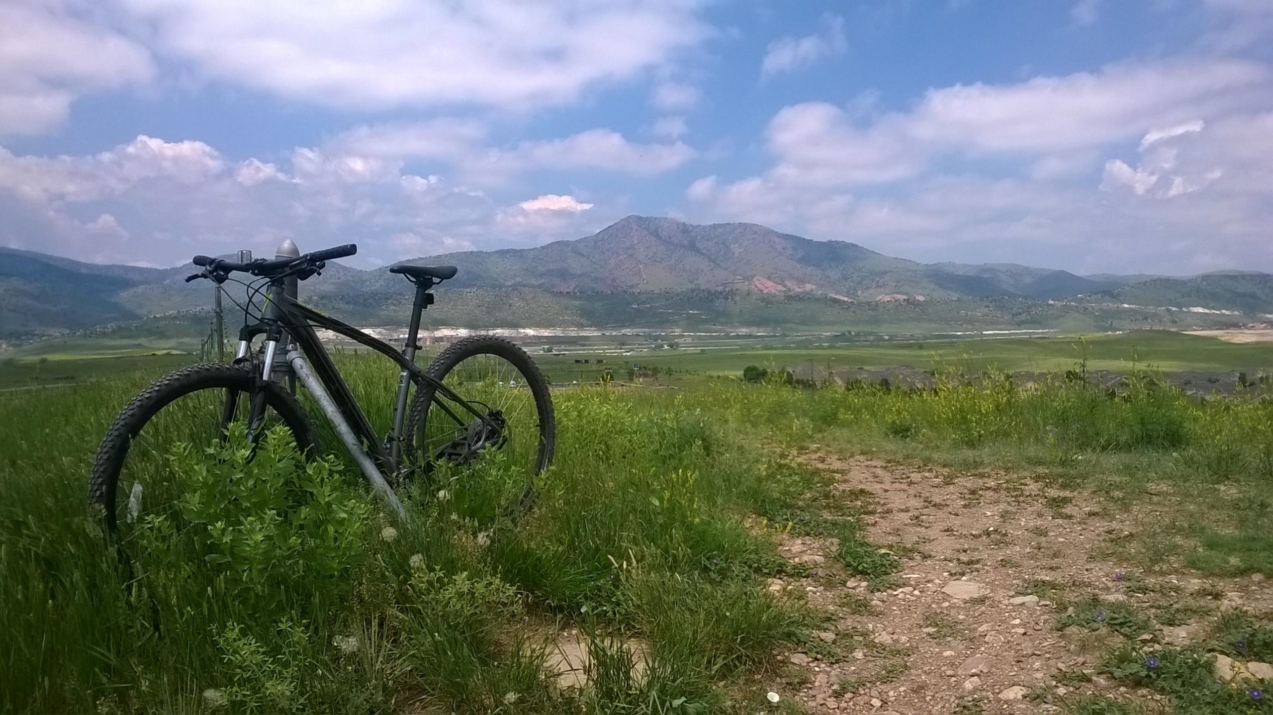 A mountain bike parked on a grassy hillside, overlooking a scenic view of rolling hills and mountains under a partly cloudy sky. Bear Creek Lake Park mountain bike trail.