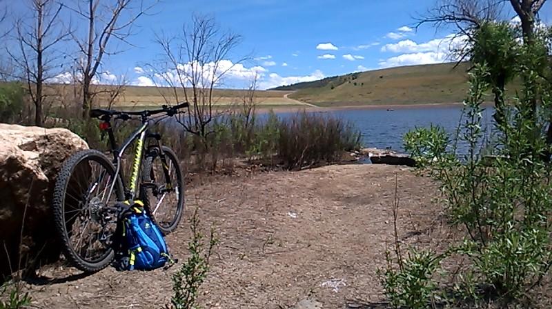 A mountain bike rests against a large rock beside a serene lake, with a blue backpack on the ground nearby. The background features a clear blue sky with scattered clouds and rolling hills. Green shrubs and trees frame the scene, creating a peaceful outdoor setting. Bear Creek Lake Park mountain bike trail.