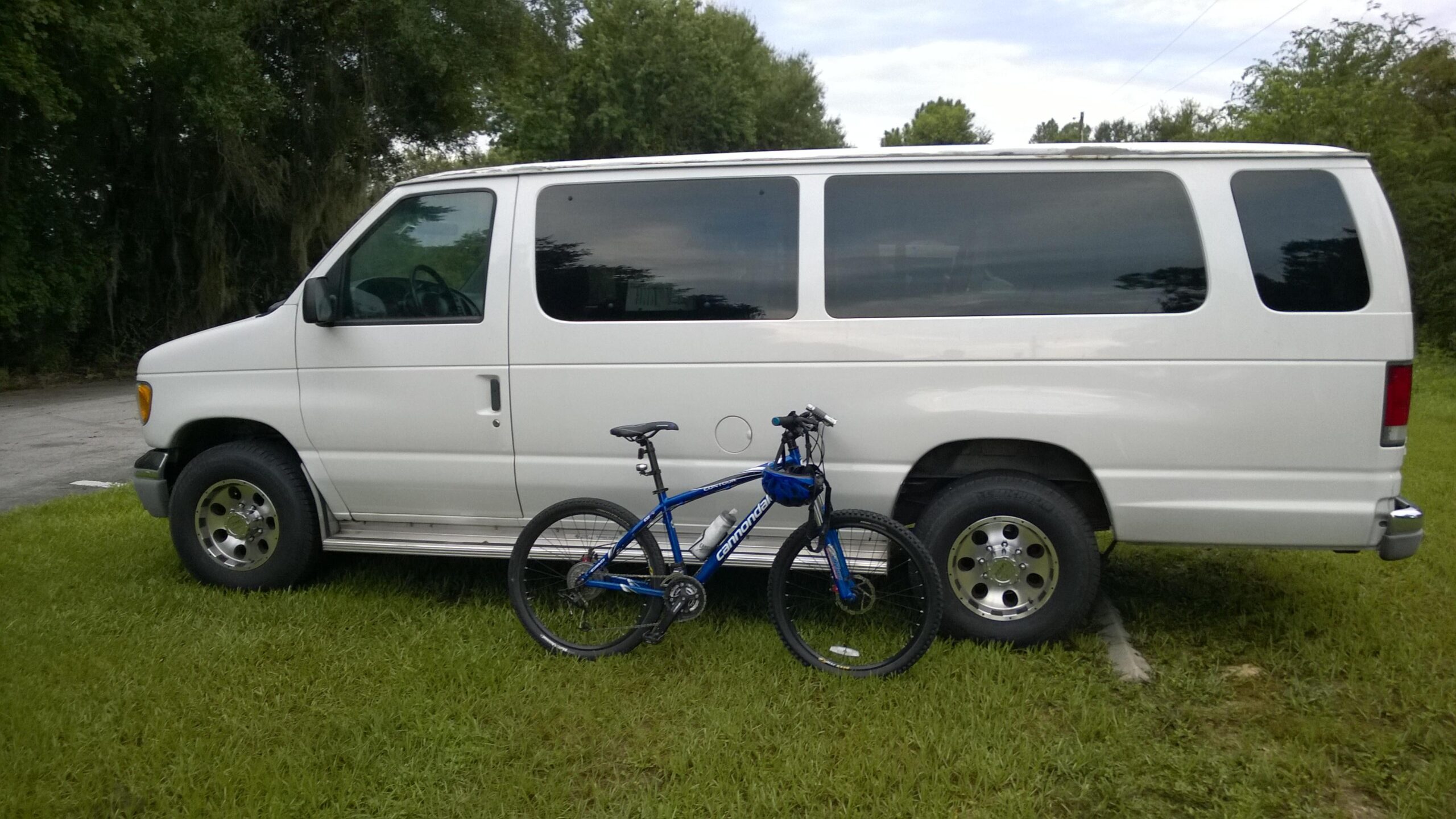Cannondale F7: A white van parked on grass next to a blue mountain bike, with trees in the background and a cloudy sky above.