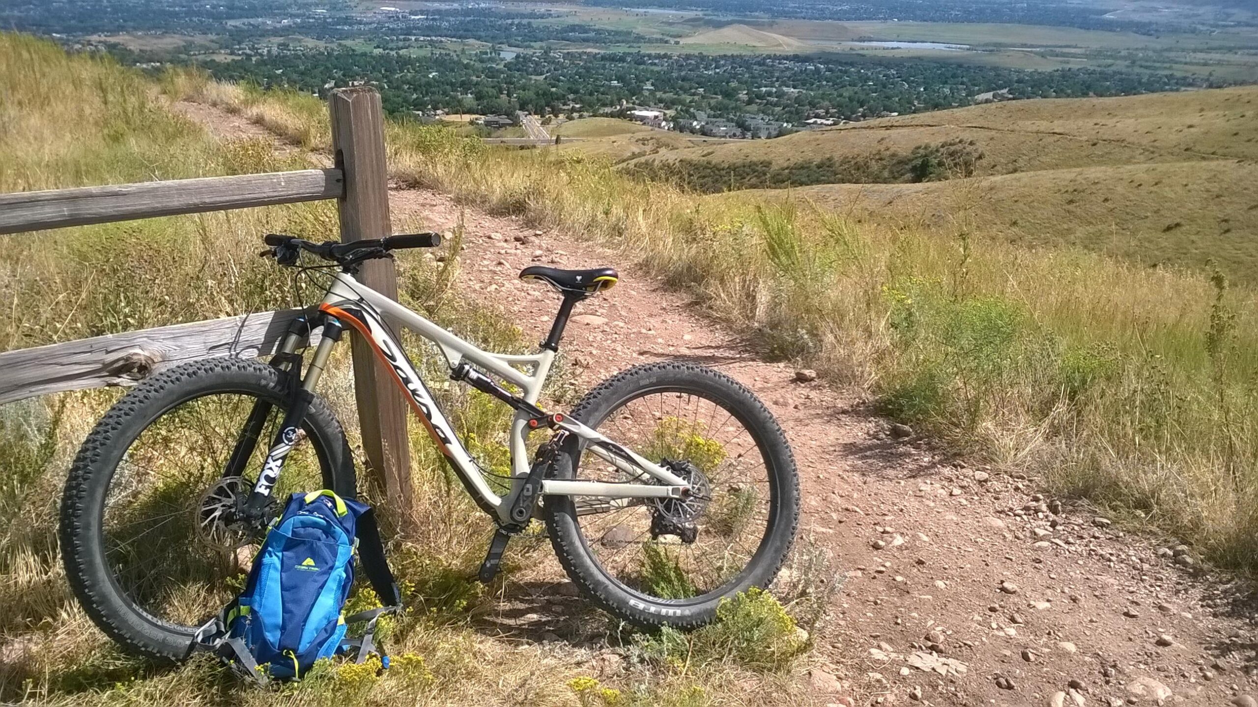 A mountain bike leaning against a wooden fence on a dirt trail, with a scenic view of a green valley and distant hills in the background. A blue backpack is placed on the ground next to the bike, surrounded by tall grass and wildflowers. Green Mountain mountain bike trail.