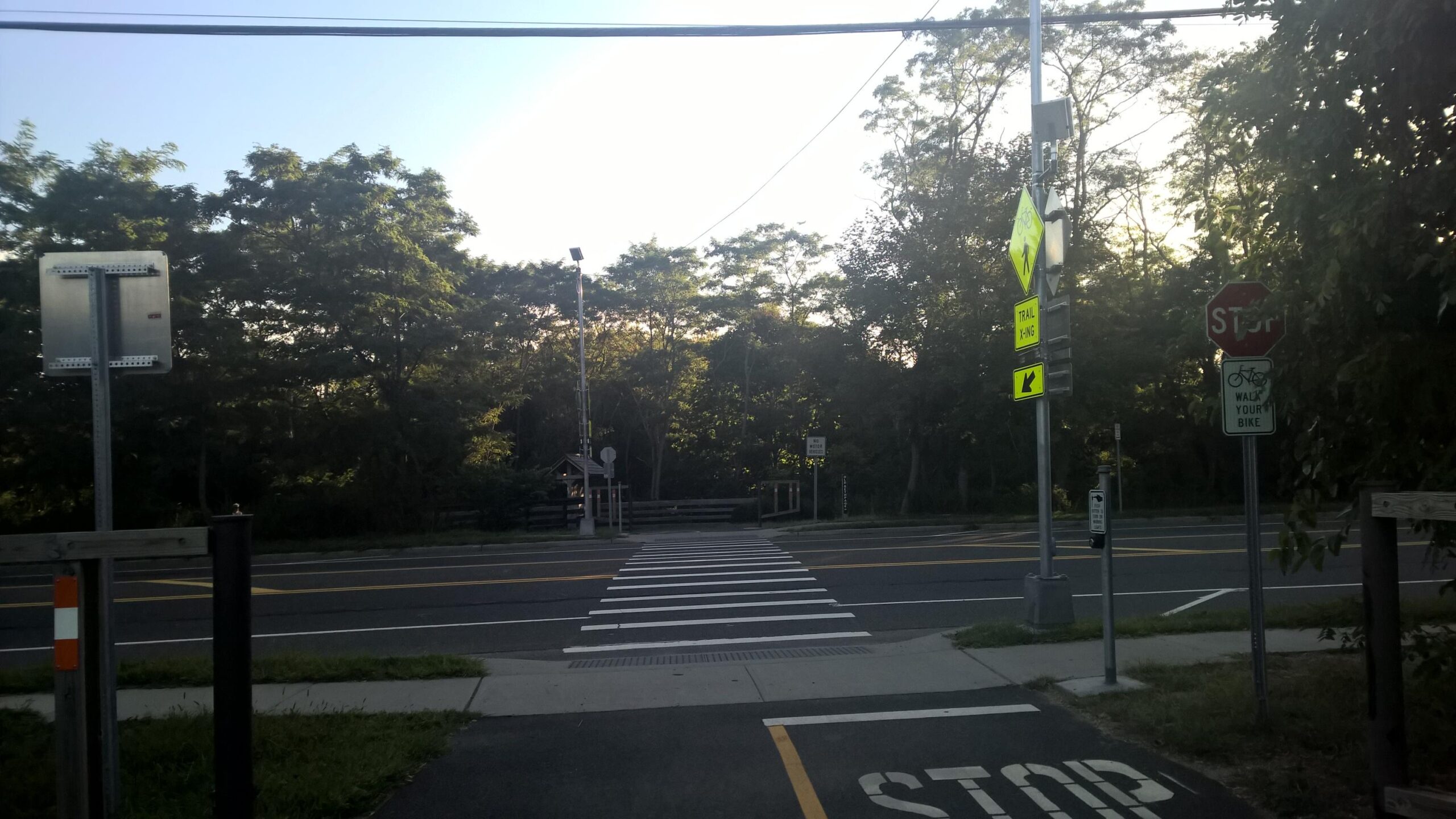 A view of a crosswalk at an intersection, with several traffic signs including stop signs and pedestrian signals. The scene is framed by trees, and a clear sky is visible in the background. The road is marked with bike and crosswalk indicators, suggesting it’s a shared space for vehicles and pedestrians. Trail View State Park mountain bike trail.