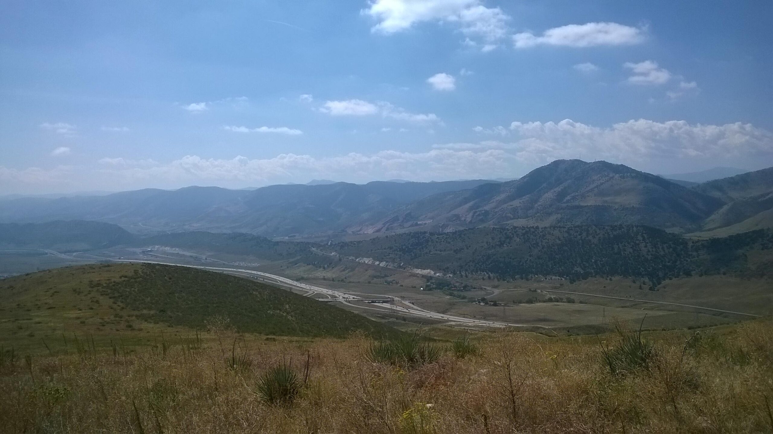 A panoramic view of rolling green hills and mountains under a blue sky with scattered clouds. A highway winds through the valley below, surrounded by lush landscapes and distant peaks. The scene captures the beauty of nature and open spaces. Green Mountain mountain bike trail.