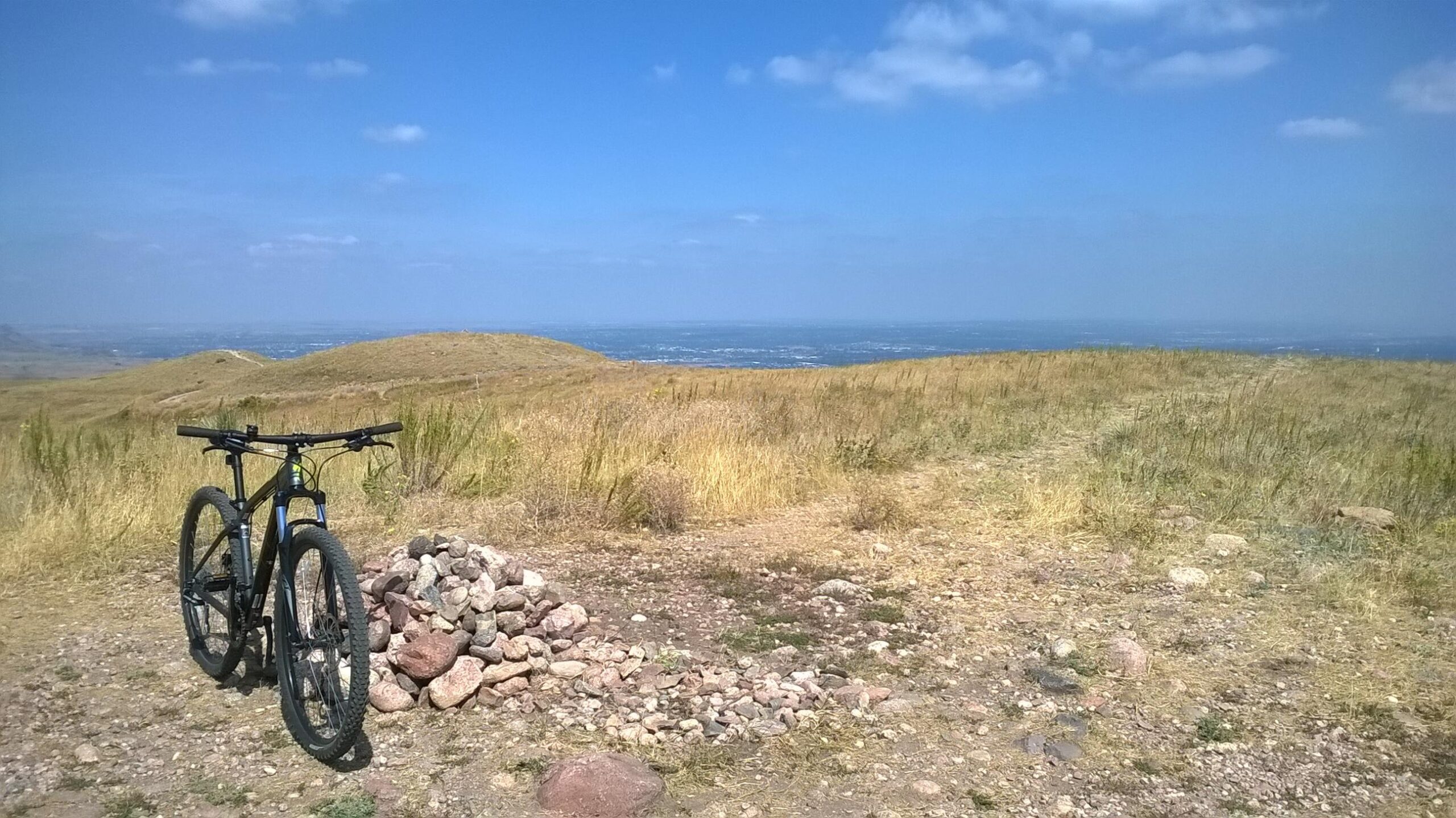 A mountain bike resting on uneven, rocky ground with a small pile of stones nearby, overlooking a vast landscape of rolling hills and distant views under a blue sky with scattered clouds. Green Mountain mountain bike trail.
