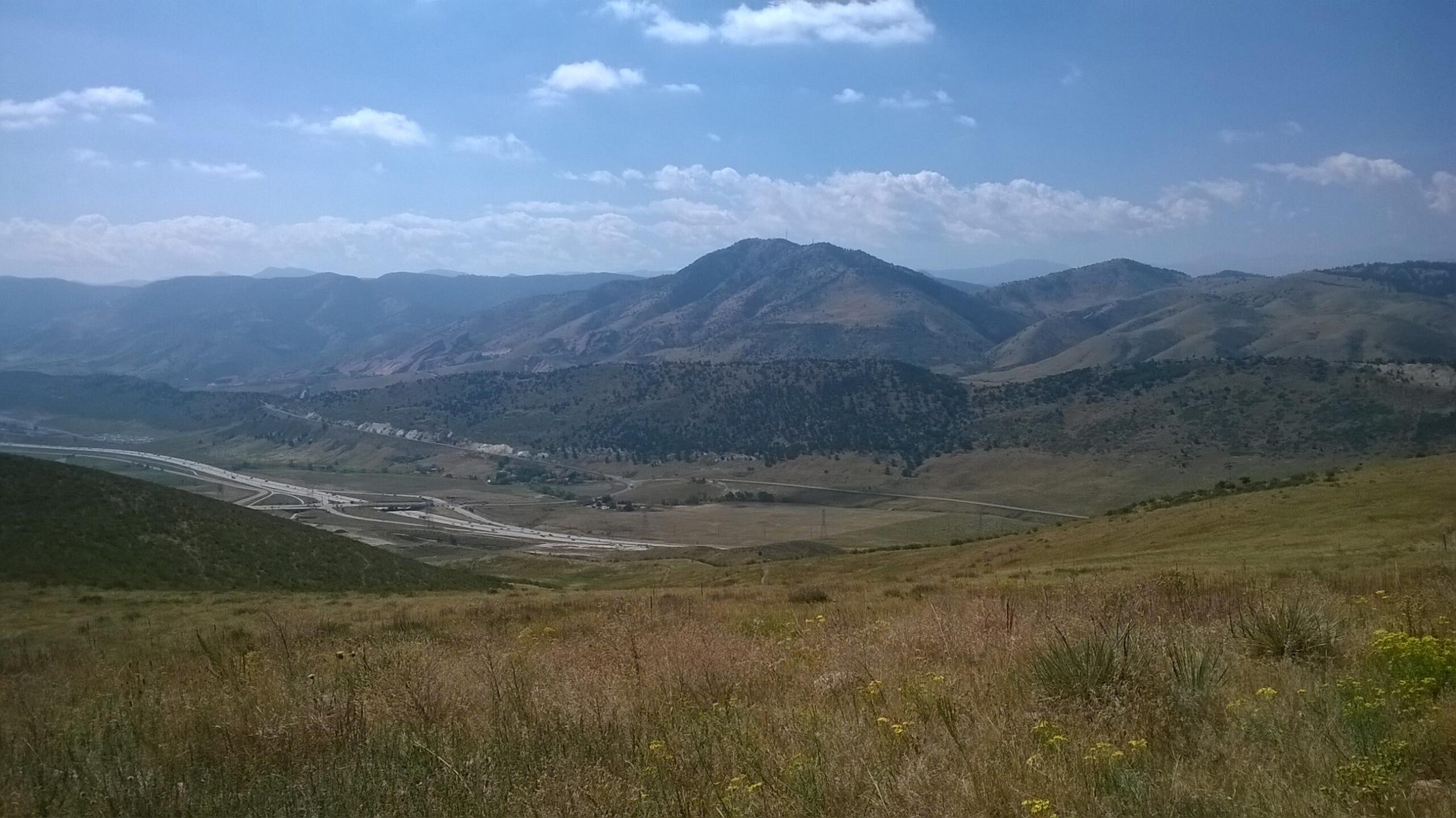 A panoramic view of rolling hills and mountains under a blue sky with fluffy white clouds. A highway runs through the valley below, surrounded by greenery and patches of wildflowers in the foreground. The landscape captures a peaceful natural setting, showcasing the beauty of the outdoors. Green Mountain mountain bike trail.