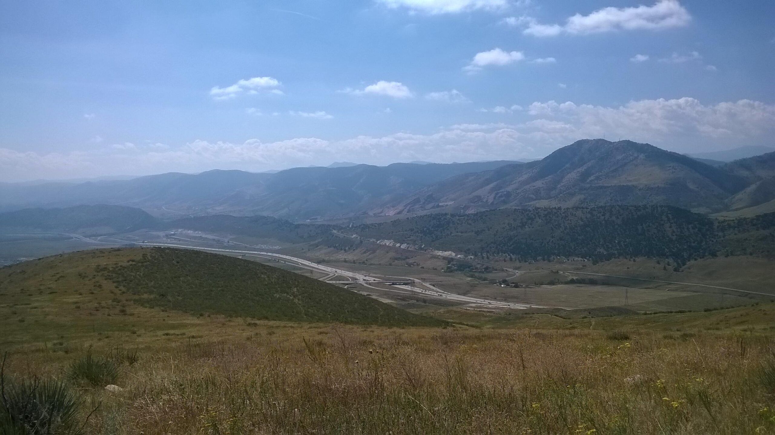 A scenic view of rolling hills and mountains under a bright blue sky with scattered clouds. A winding road is visible in the valley below, surrounded by grassy terrain and patches of greenery. The landscape features a mix of grassy hills and rugged mountains in the background, creating a serene natural environment. Green Mountain mountain bike trail.