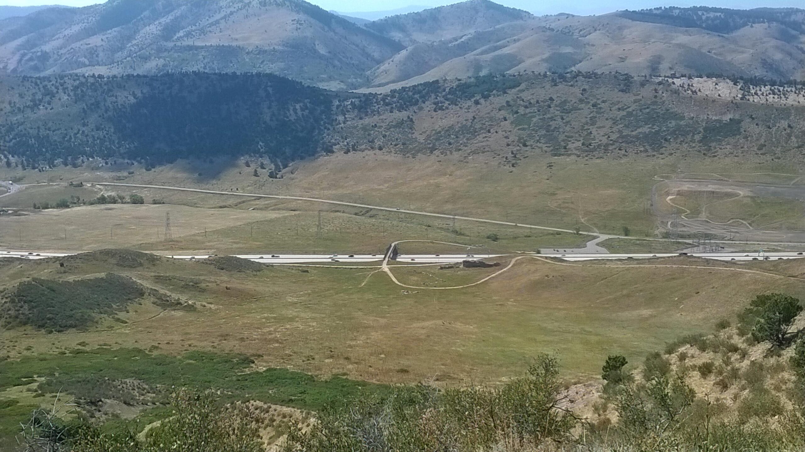 A panoramic view of rolling hills and mountains, showcasing a highway that winds through the landscape. The foreground features grassy plains and shrubs, while the background includes rugged mountain ranges under a clear blue sky. Green Mountain mountain bike trail.