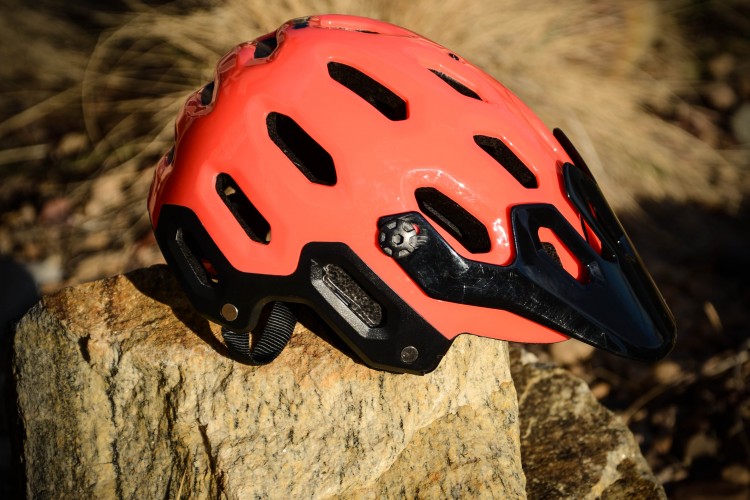 A vibrant coral helmet with black accents resting on a rock, set against a blurred natural background. The helmet features ventilation holes and a sturdy design, suitable for cycling or outdoor activities.