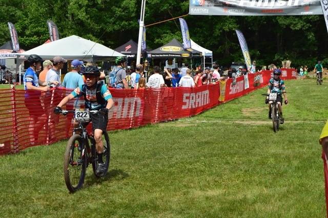 Two young cyclists are approaching the finish line at a mountain biking event, with one rider in the foreground wearing a helmet and jersey number 822. They are riding on a grassy area near a red SRAM banner, and spectators can be seen in the background, enjoying the event. Tents and flags indicate an active racing atmosphere on a sunny day.