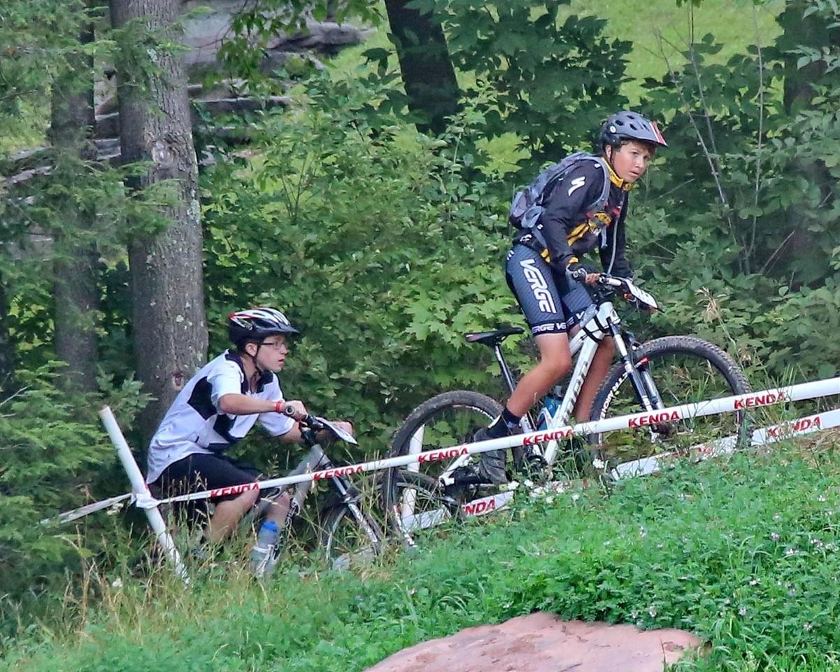 Two young cyclists navigate a lush, green trail. One cyclist, wearing a black and yellow jersey, is ascendant on a mountain bike, while the other, dressed in a white and black top, is in the background, pushing their bike uphill. The backdrop features trees and a grassy area, with race tape marking the course.