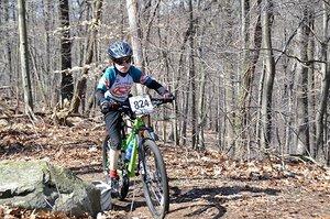A young child wearing a blue and black cycling jersey and a helmet rides a mountain bike along a dirt trail in a wooded area. The bike has a number plate labeled "824." The background features trees and fallen leaves.