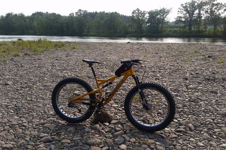 A bright orange fat bike stands on a rocky shore beside a calm river, surrounded by greenery and trees in the background under a clear sky.