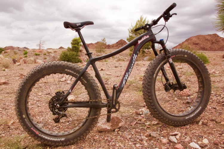 A black fat tire mountain bike is positioned on rocky terrain, surrounded by sparse vegetation and desert-like scenery. The bike features wide tires designed for off-road durability and a sleek frame with visible branding. The sky is overcast, suggesting a cloudy day.