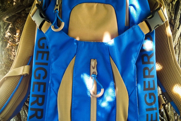 A blue and tan Geigerig RIG 650 backpack hanging against a tree, featuring various pockets and straps for storage and comfort. The backpack has a prominent logo on the front and is designed for outdoor activities.