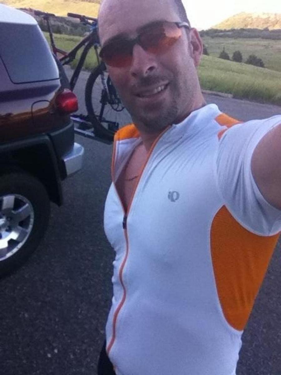 Specialized S-Works Stumpjumper FSR: A man in a white cycling jersey with orange accents is smiling at the camera while standing next to a parked vehicle. A mountain bike is mounted on the back of the car, and a scenic landscape with greenery and mountains is visible in the background. The sun appears to be shining, creating a bright and cheerful atmosphere.