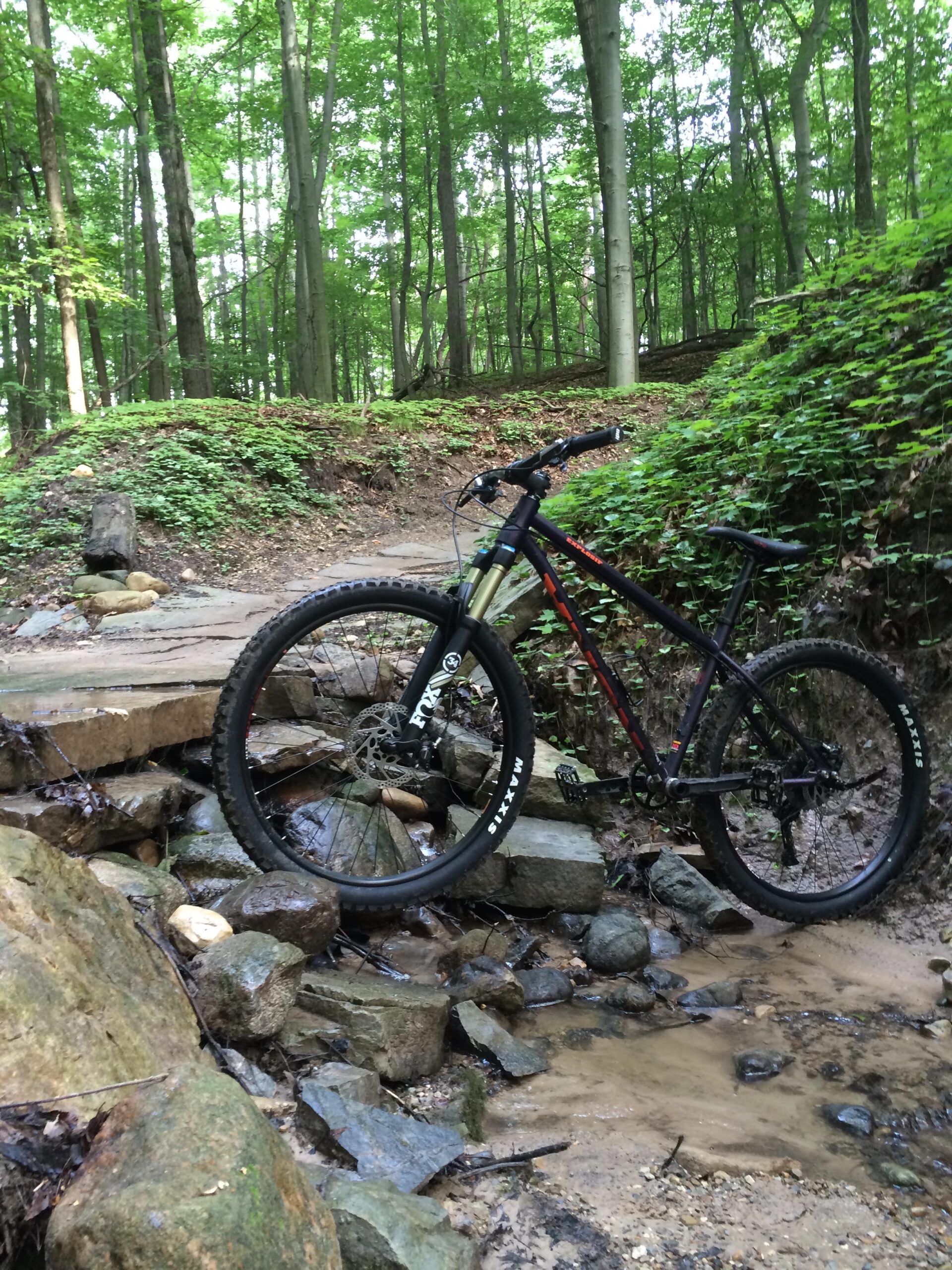 Kona EXPLOSIF: A mountain bike is positioned on a rocky path crossing a small stream in a lush green forest. The surrounding trees are tall and provide a vibrant canopy of leaves, with patches of sunlight filtering through. The scene captures a natural, rugged trail ideal for biking or outdoor adventures.
