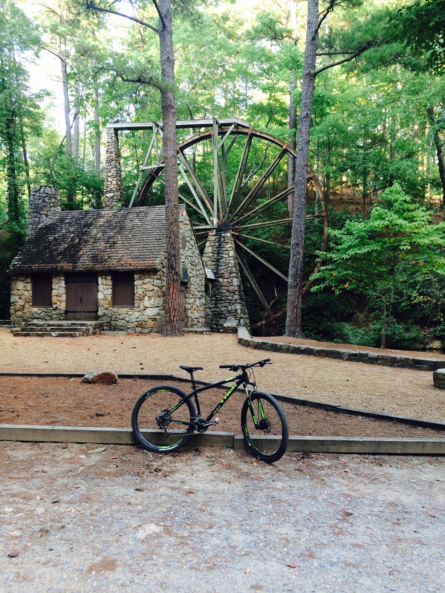 Trek X-Caliber: A mountain bike is parked on a gravel path in front of a stone structure with a thatched roof, set in a lush green forest. A large, wooden water wheel is visible behind the building, surrounded by trees.