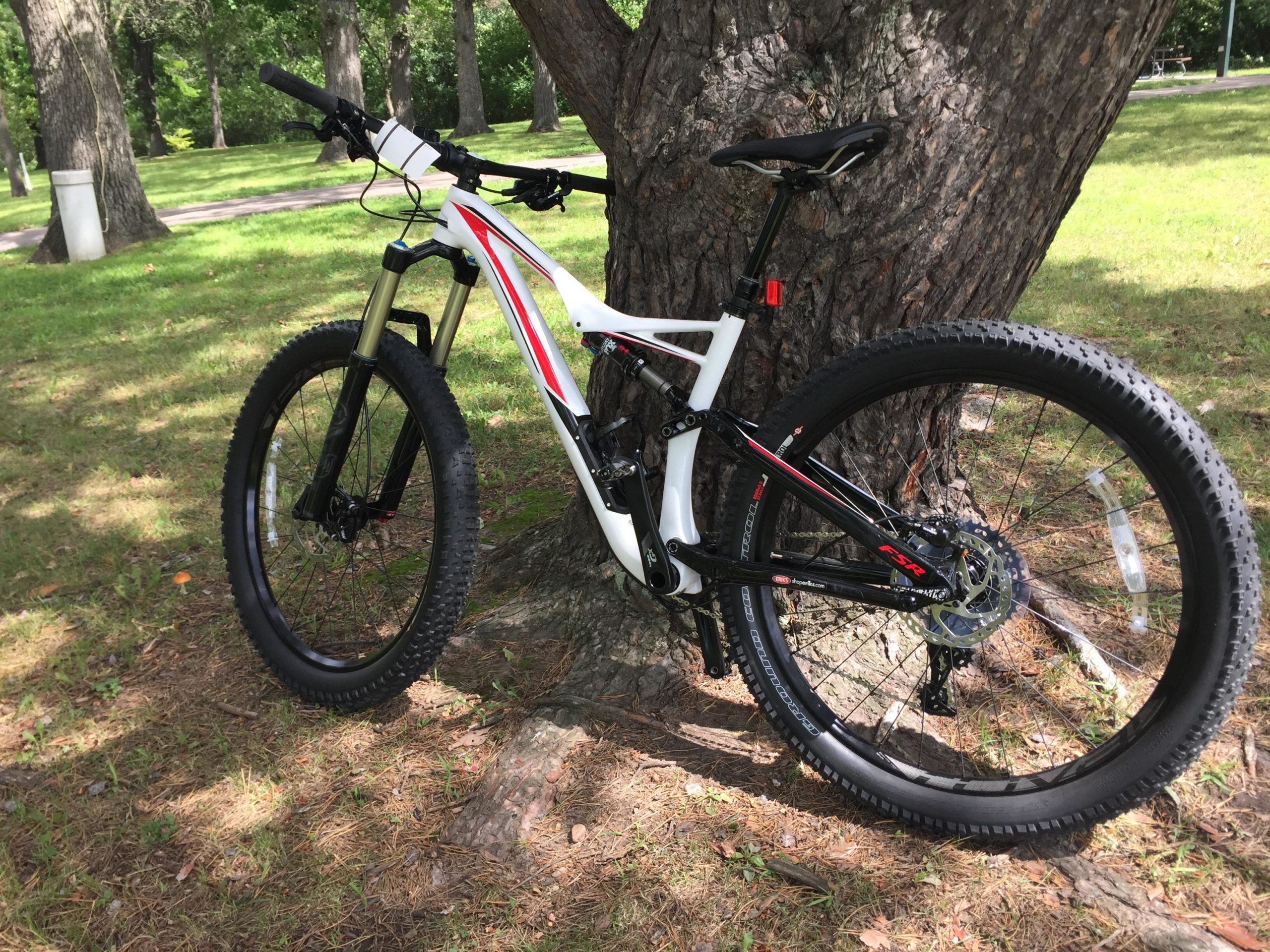 A mountain bike with a white and red frame, positioned against a tree in a grassy park setting. The bike features thick tires and a front suspension fork, indicating it is designed for off-road use. Sunlight filters through the trees, creating a natural and relaxed atmosphere. Mammoth Trail mountain bike trail.