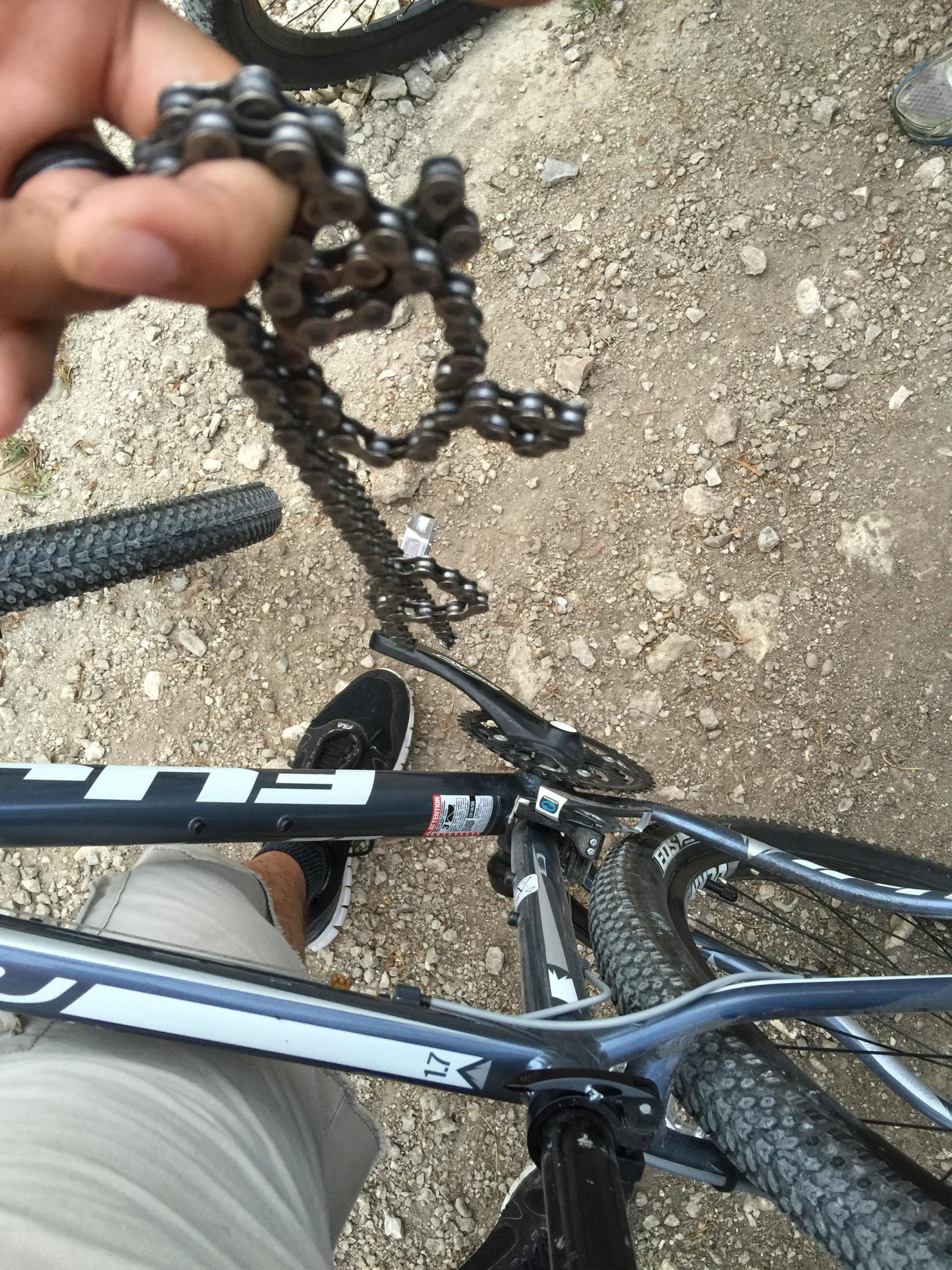 A close-up view of a hand holding a detached mountain bike chain above a bike frame with gravel and dirt underneath. The image also shows part of a foot wearing a black sneaker and the bike’s tires in the background. Dana Peak mountain bike trail.
