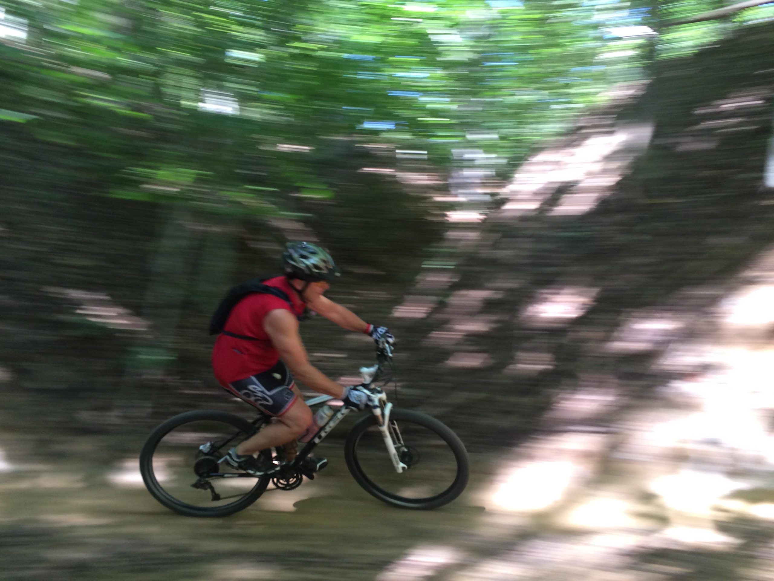 A cyclist in a red shirt rides swiftly along a wooded trail, surrounded by blurred greenery, suggesting high speed and motion. Overmountain Victory Trail mountain bike trail.