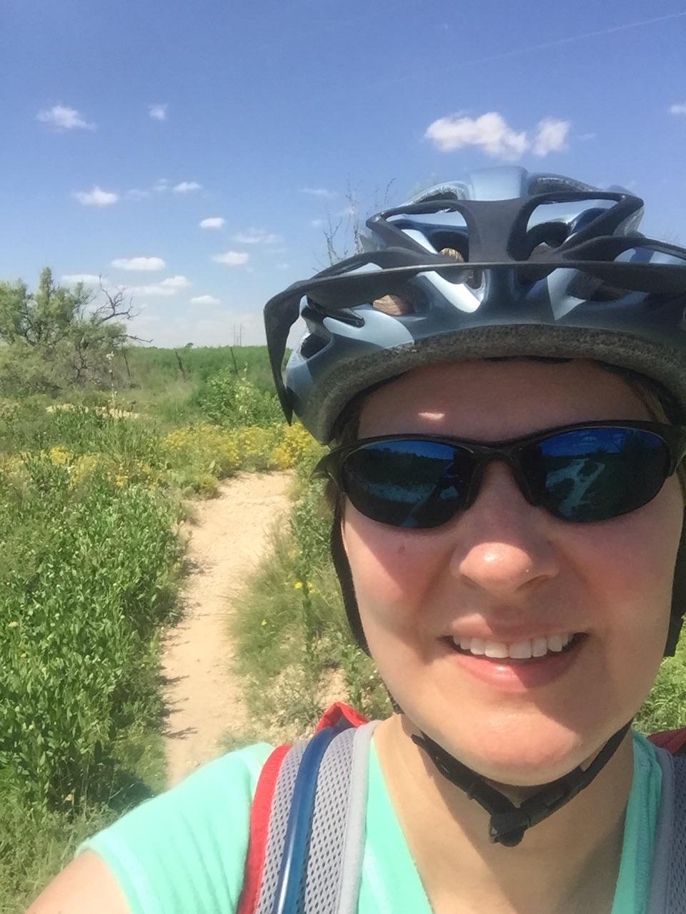 Santa Cruz Tallboy Carbon: A person wearing sunglasses and a light blue helmet smiles for a selfie while biking on a sunny day. Green vegetation and wildflowers line a dirt path in the background, under a blue sky with a few clouds.