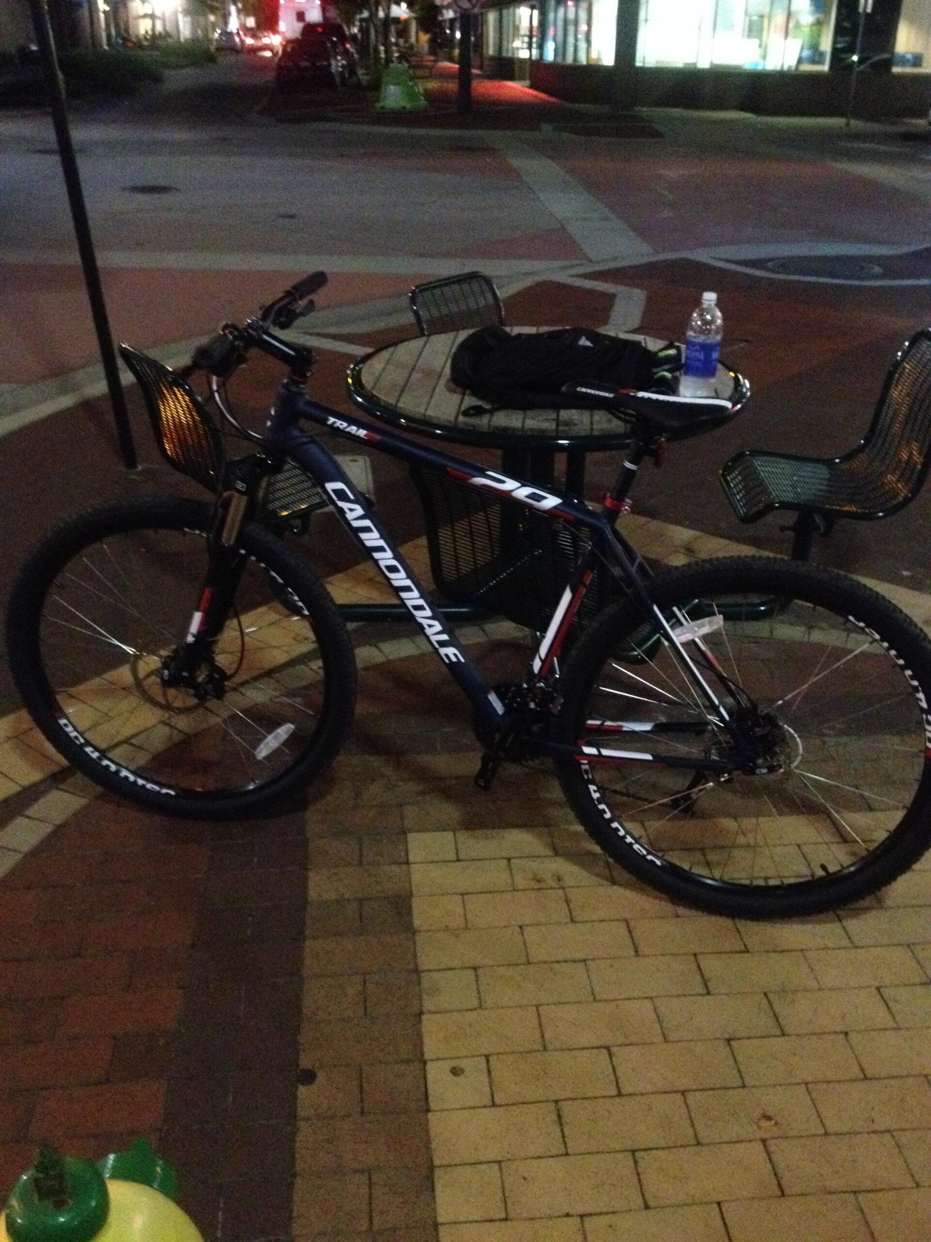 Cannondale Trail 5: A Cannondale mountain bike is parked next to a green and black table and chairs in a well-lit outdoor area at night. A water bottle and a black garment are resting on the table. The ground is made of patterned bricks.