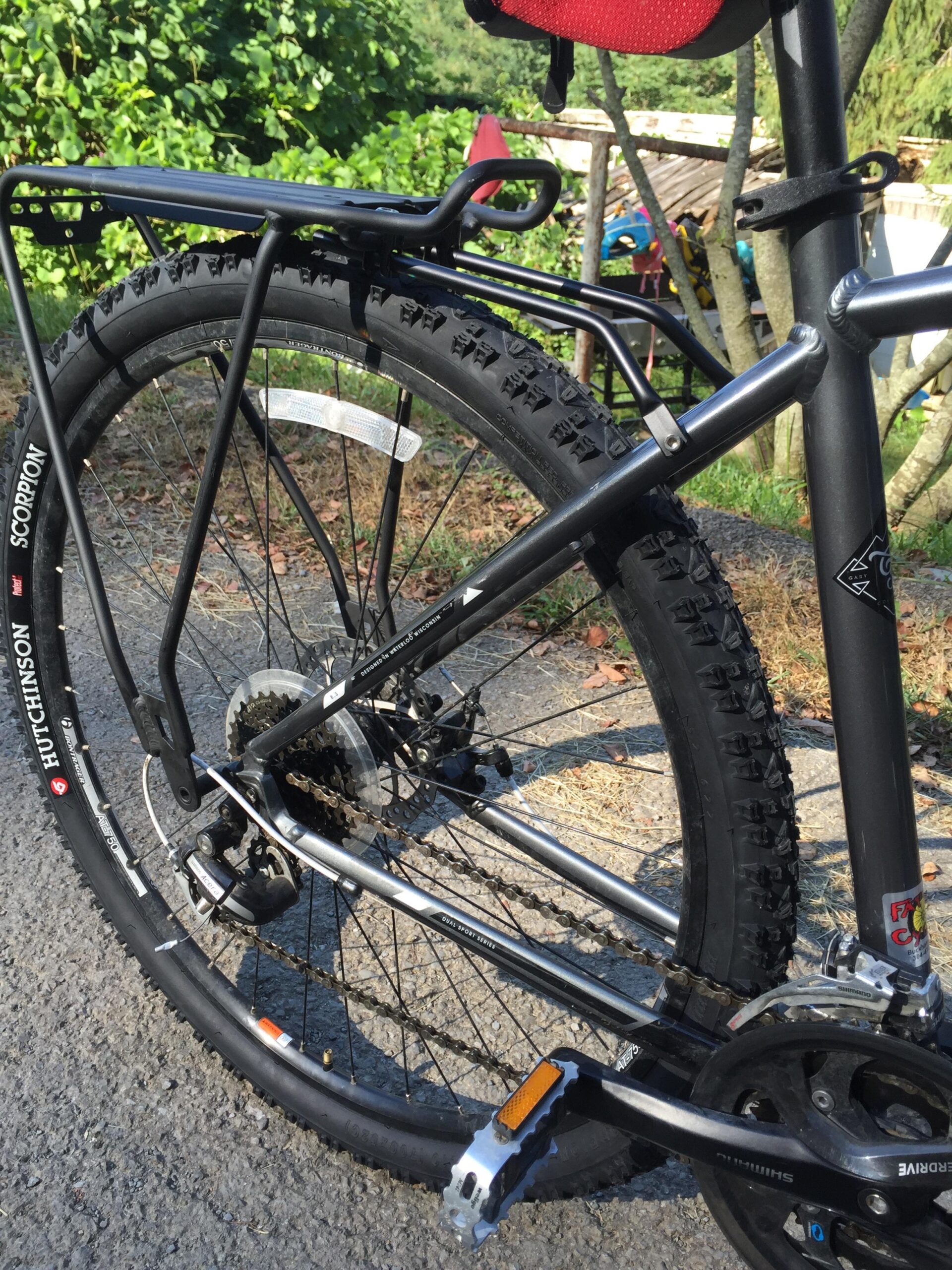 Hutchinson Scorpion: Close-up view of the rear wheel and bike components, featuring a rugged tire, a rear rack, and a chain mechanism. The background includes greenery and a hint of other outdoor structures.