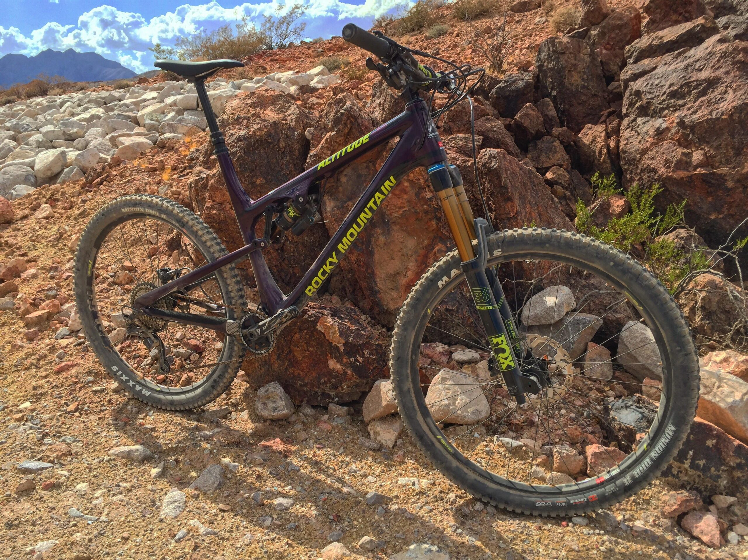 Mountain bike leaning against rocks in a rugged outdoor environment, with a backdrop of blue skies and scattered clouds. The bike features a purple frame with "Rocky Mountain" branding, equipped with quality tires and suspension for off-road riding. Bootleg Canyon mountain bike trail.