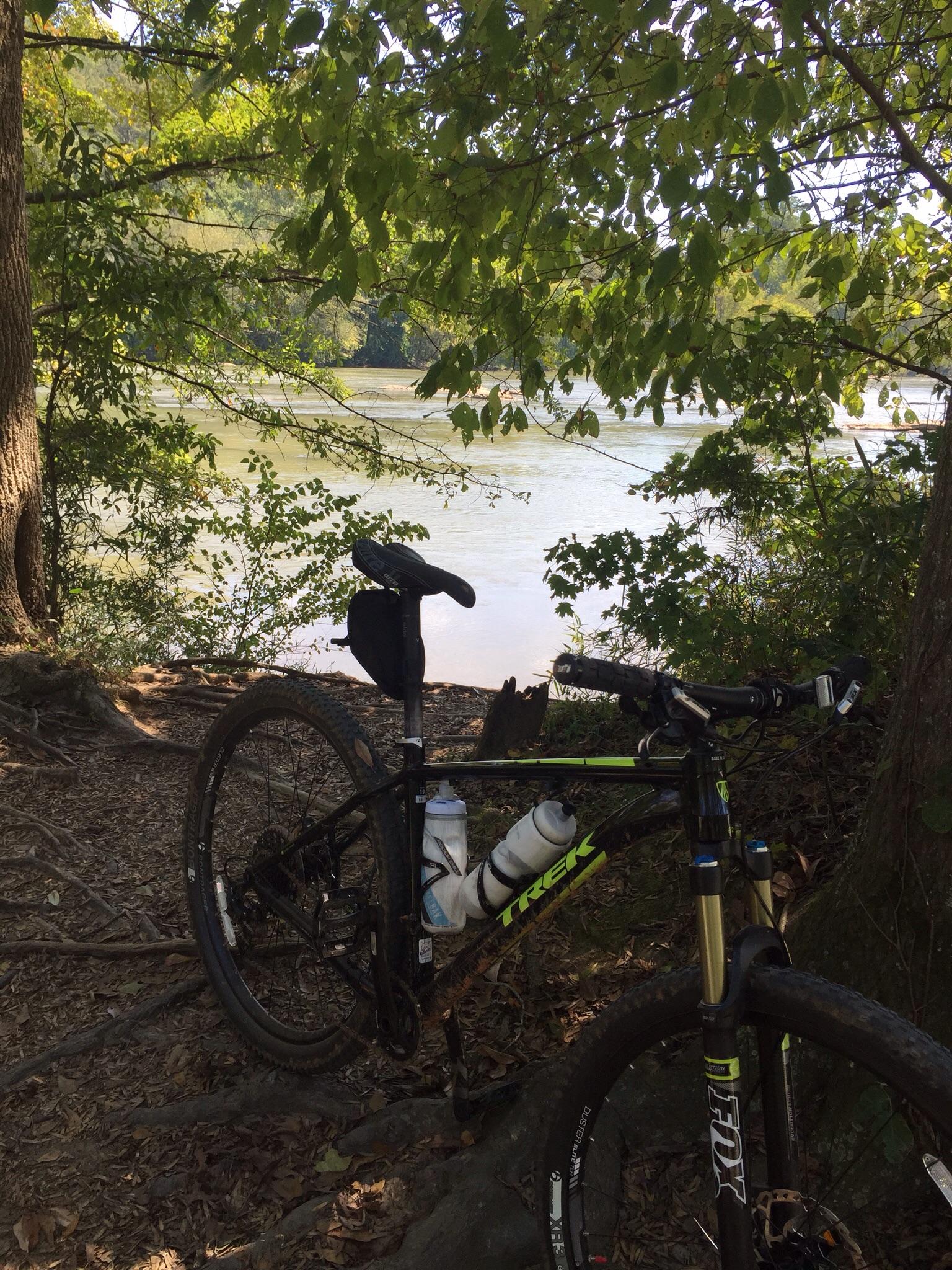 Trek Stache 8: A mountain bike parked near a river, surrounded by trees and lush greenery. The bike has a water bottle attached to its frame, and the scene captures a tranquil outdoor setting.