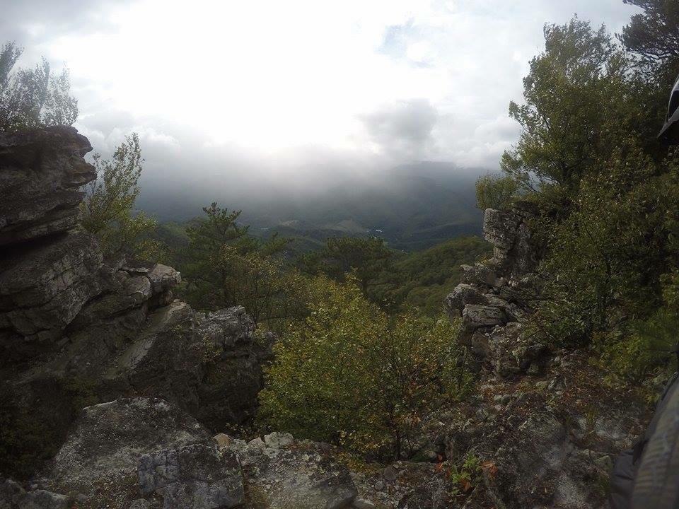 A panoramic view of a mountainous landscape with rocky outcrops in the foreground. The scene is partly cloudy, with mist hovering over the distant hills, and lush greenery surrounding the area, creating a serene and natural atmosphere. North Fork Mountain Trail mountain bike trail.