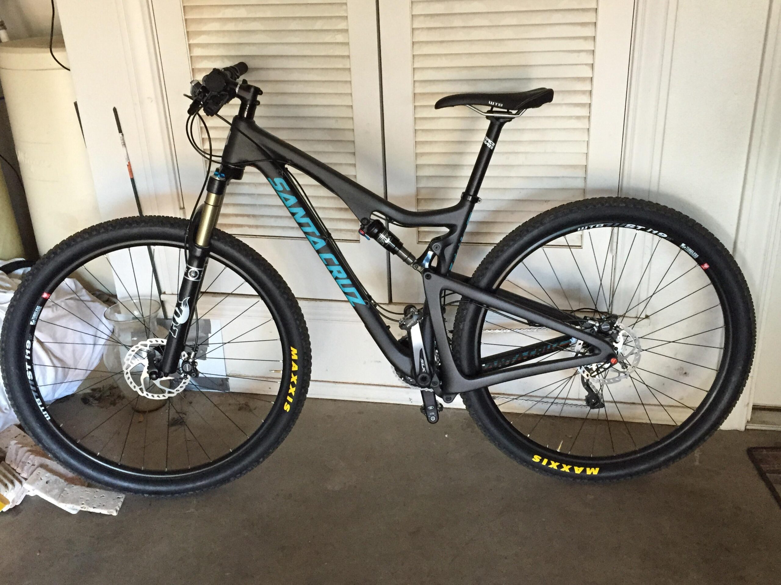 Santa Cruz Tallboy Carbon: A black Santa Cruz mountain bike with blue accents, featuring front suspension, wide tires with Maxxis branding, and disc brakes. The bike is positioned in a garage setting with a closed shutter in the background.