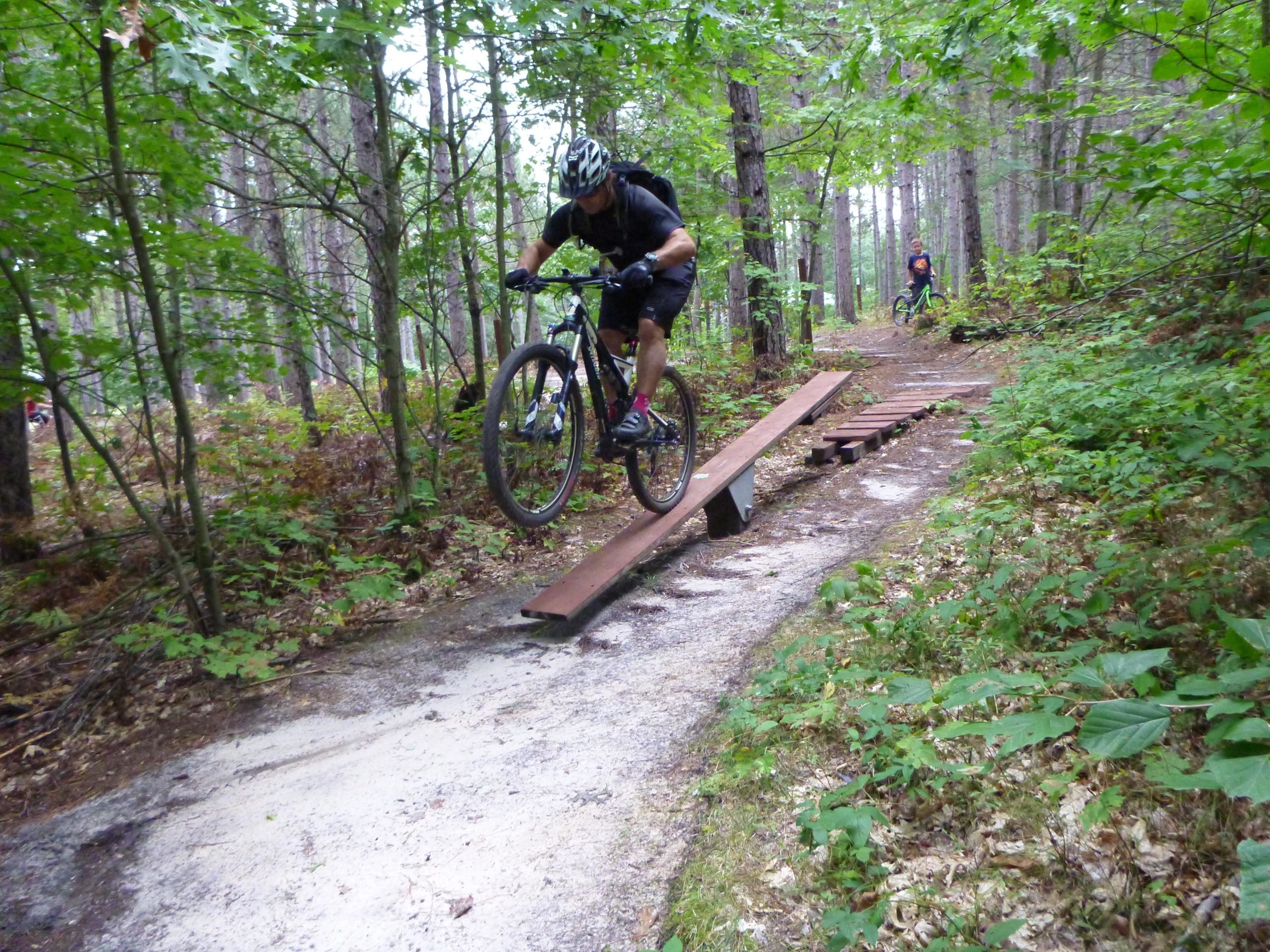 A mountain biker jumps off a wooden ramp on a forest trail, surrounded by trees and greenery. Another cyclist can be seen in the background, navigating the same path. Levis Mounds mountain bike trail.