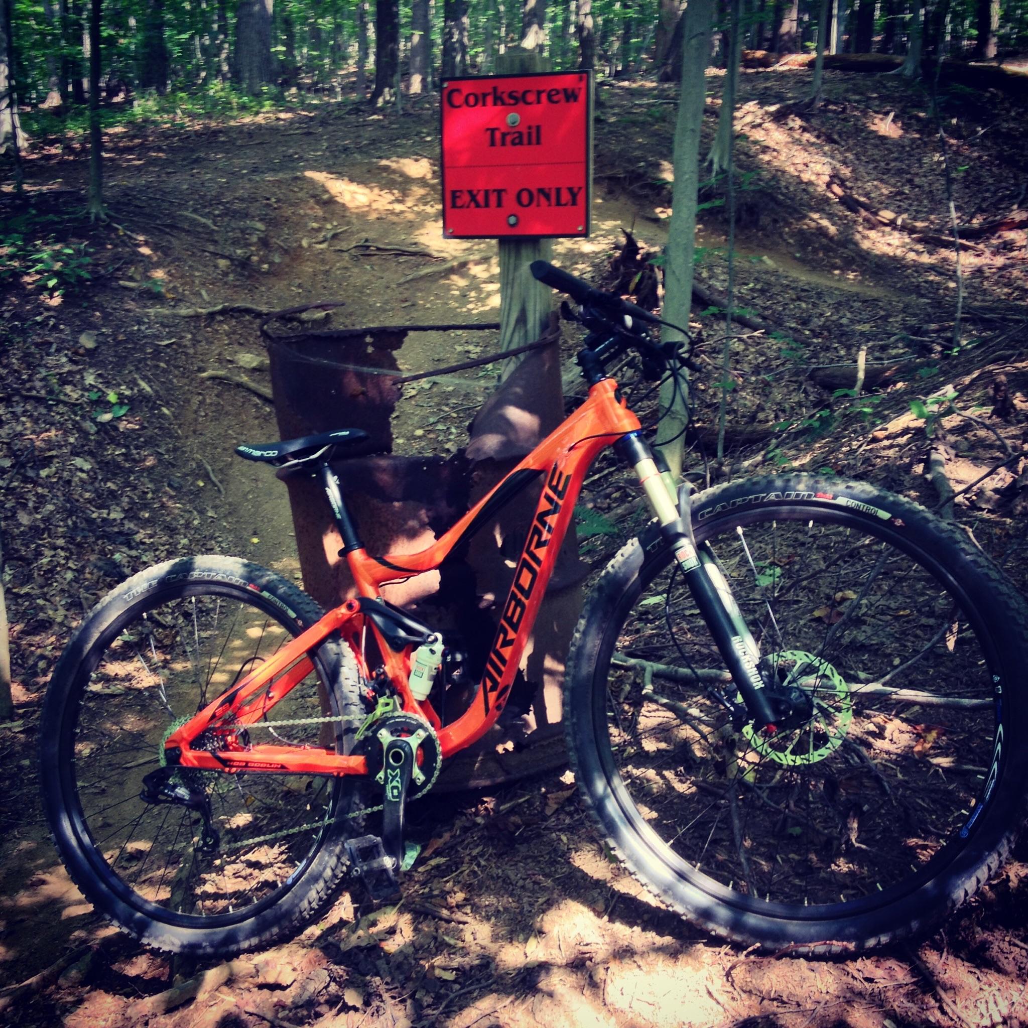 Airborne Hobgoblin: A mountain bike with an orange frame is parked next to a red sign that reads "Corkscrew Trail - EXIT ONLY." The background features a wooded area with trees and leaves scattered on the ground.