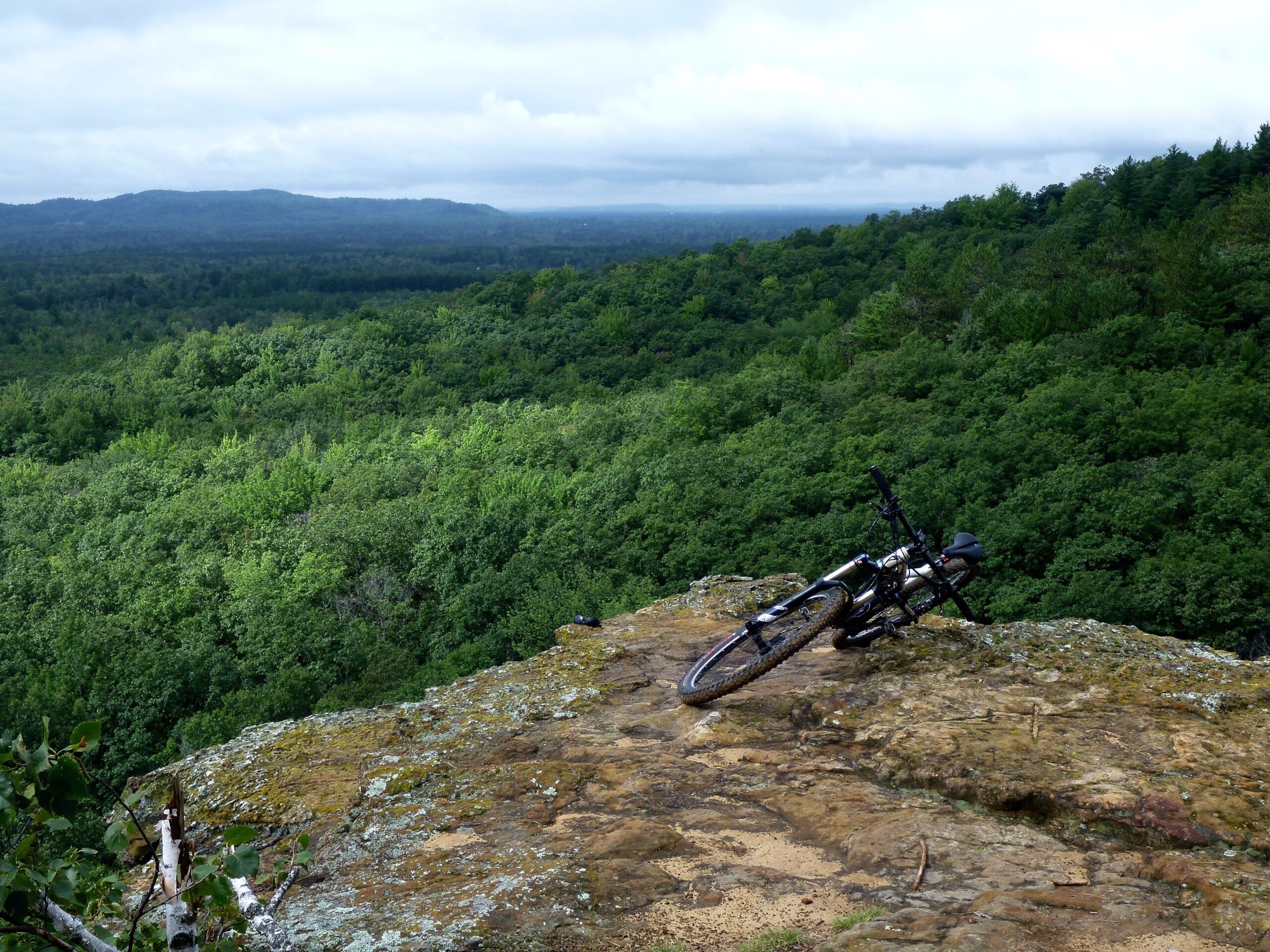 A mountain bike rests on a rocky ledge overlooking a dense, green forest and distant hills under a cloudy sky. Levis Mounds mountain bike trail.
