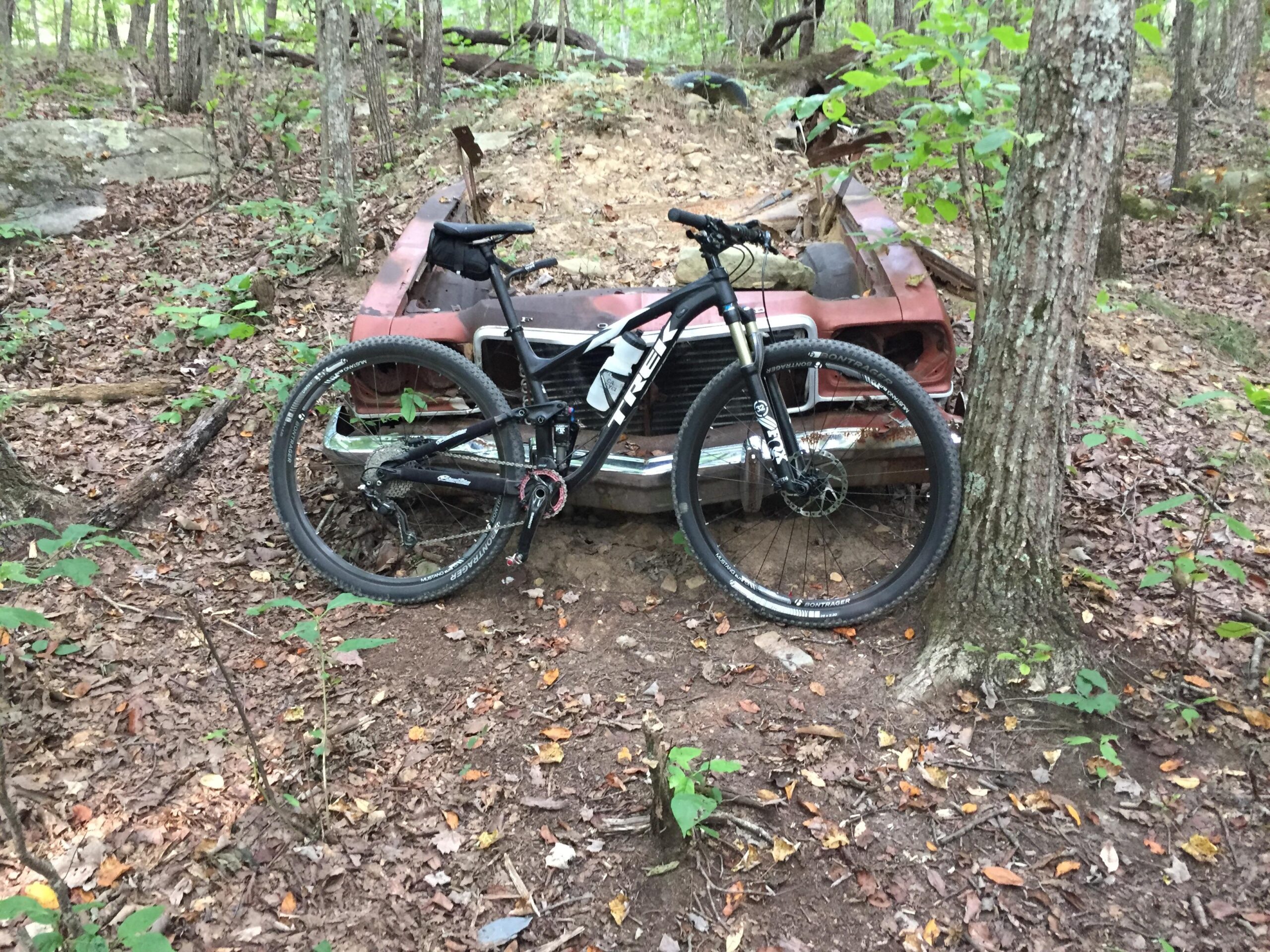 Trek Top Fuel 8: A mountain bike resting against a partially buried vintage car in a wooded area, surrounded by fallen leaves and trees. The scene captures the blend of nature and remnants of human activity in an outdoor setting.