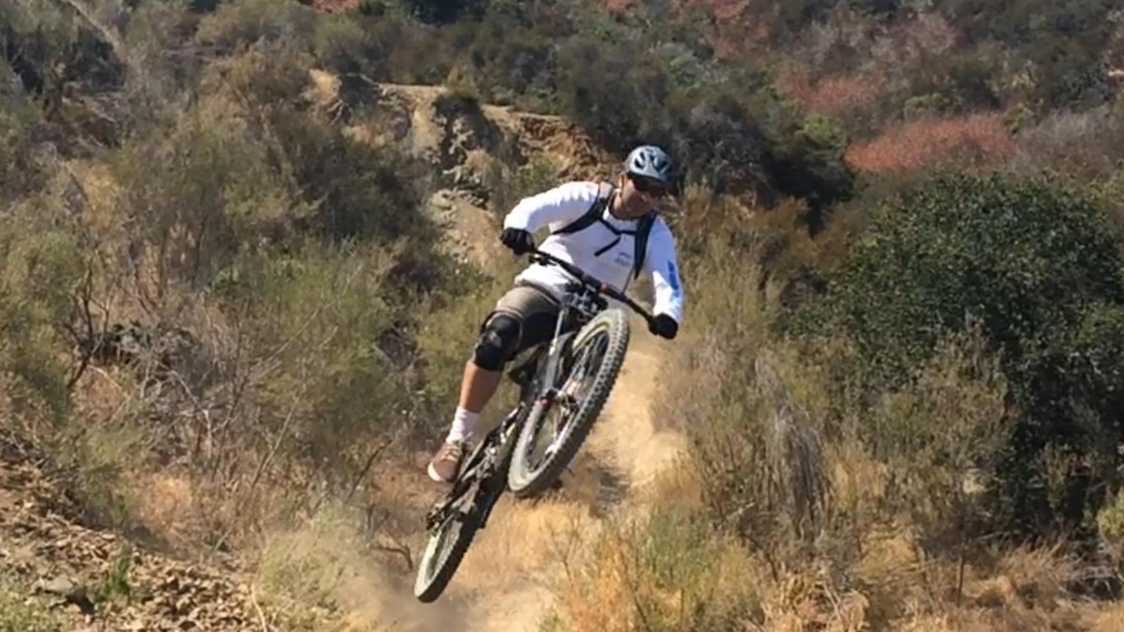 Specialized S-Works Demo 8 Carbon: A mountain biker performing a jump on a dirt trail surrounded by bushes and rocky terrain. The rider is wearing a helmet and protective gear while capturing the thrill of biking in an outdoor setting. Dust is kicked up from the trail, emphasizing the dynamic movement.