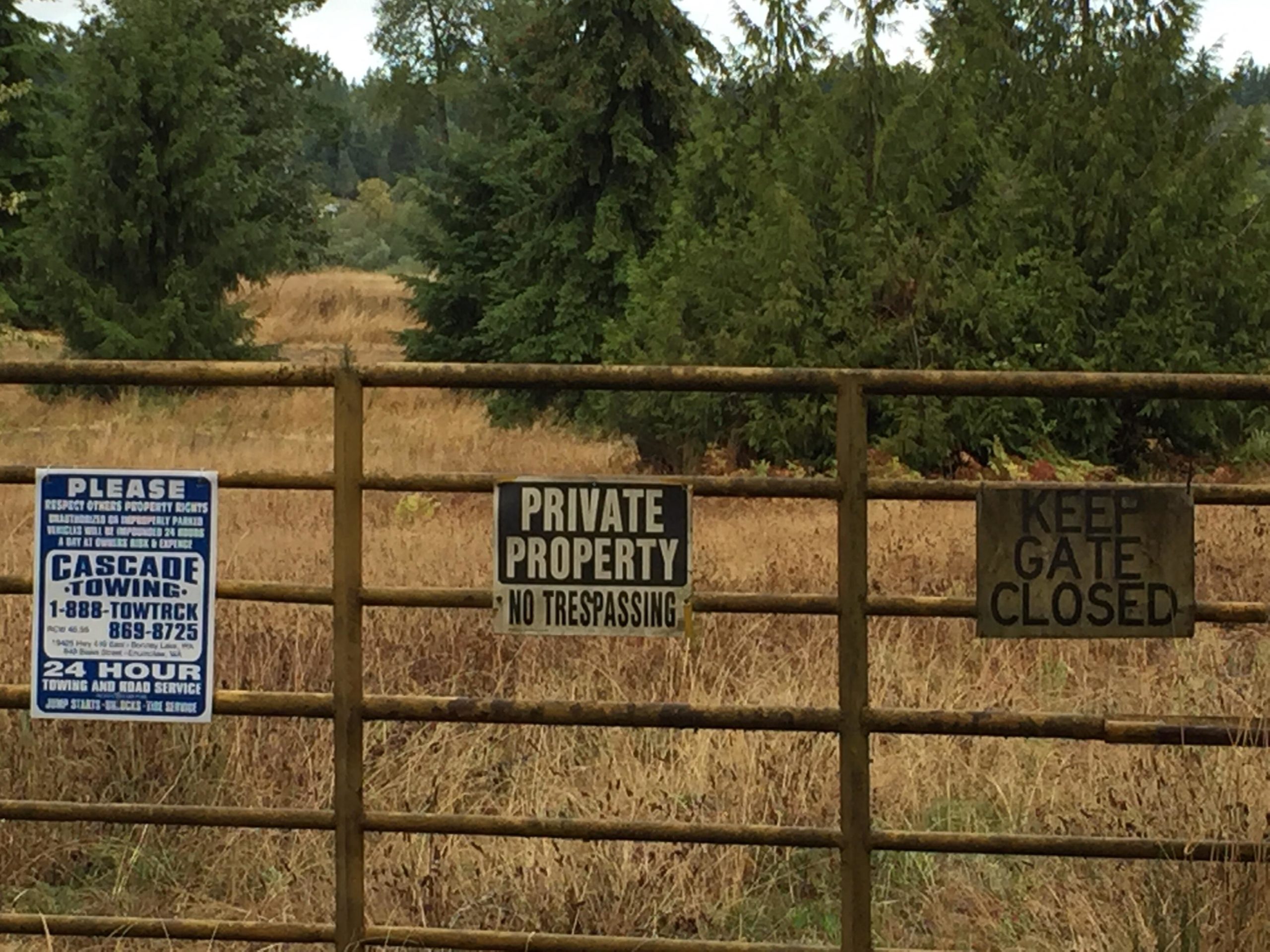 A gated entrance featuring several signs, including one that says "Private Property - No Trespassing," another that instructs to "Keep Gate Closed," and a third sign for a towing service, emphasizing respect for property rights. The surroundings include tall grass and trees, indicating a rural setting. Victor Falls mountain bike trail.