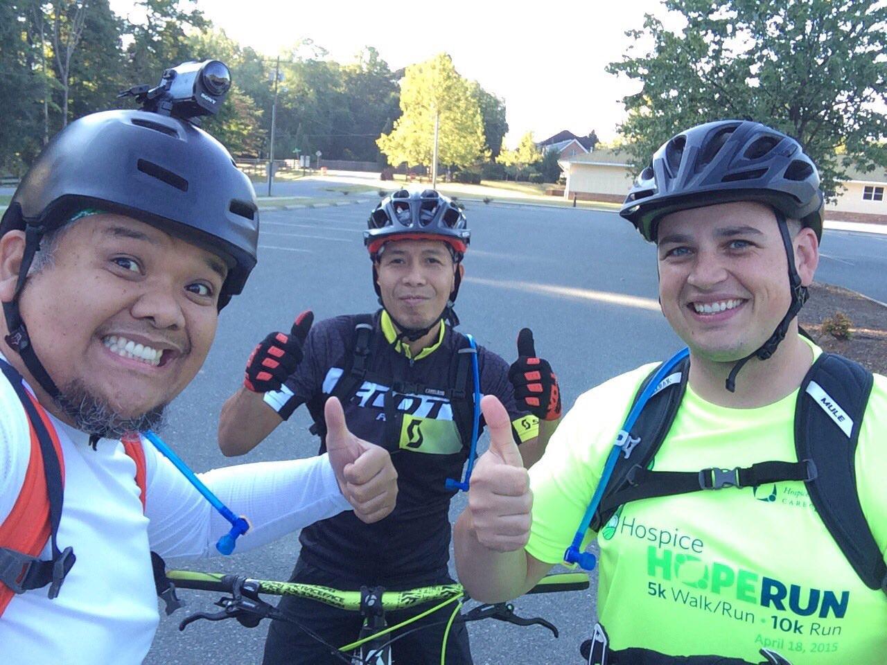 Three individuals wearing helmets and cycling gear are posing for a selfie outdoors. The first person, on the left, has a broad smile and a camera mounted on his helmet. The second person, in the middle, is giving a thumbs-up and is dressed in a dark athletic shirt with yellow accents. The third person, on the right, is also giving a thumbs-up and is wearing a bright neon green shirt that promotes a hospice run event. They all appear cheerful and ready for an outdoor biking adventure. Owls Roost (Bur-Mil Park) mountain bike trail.
