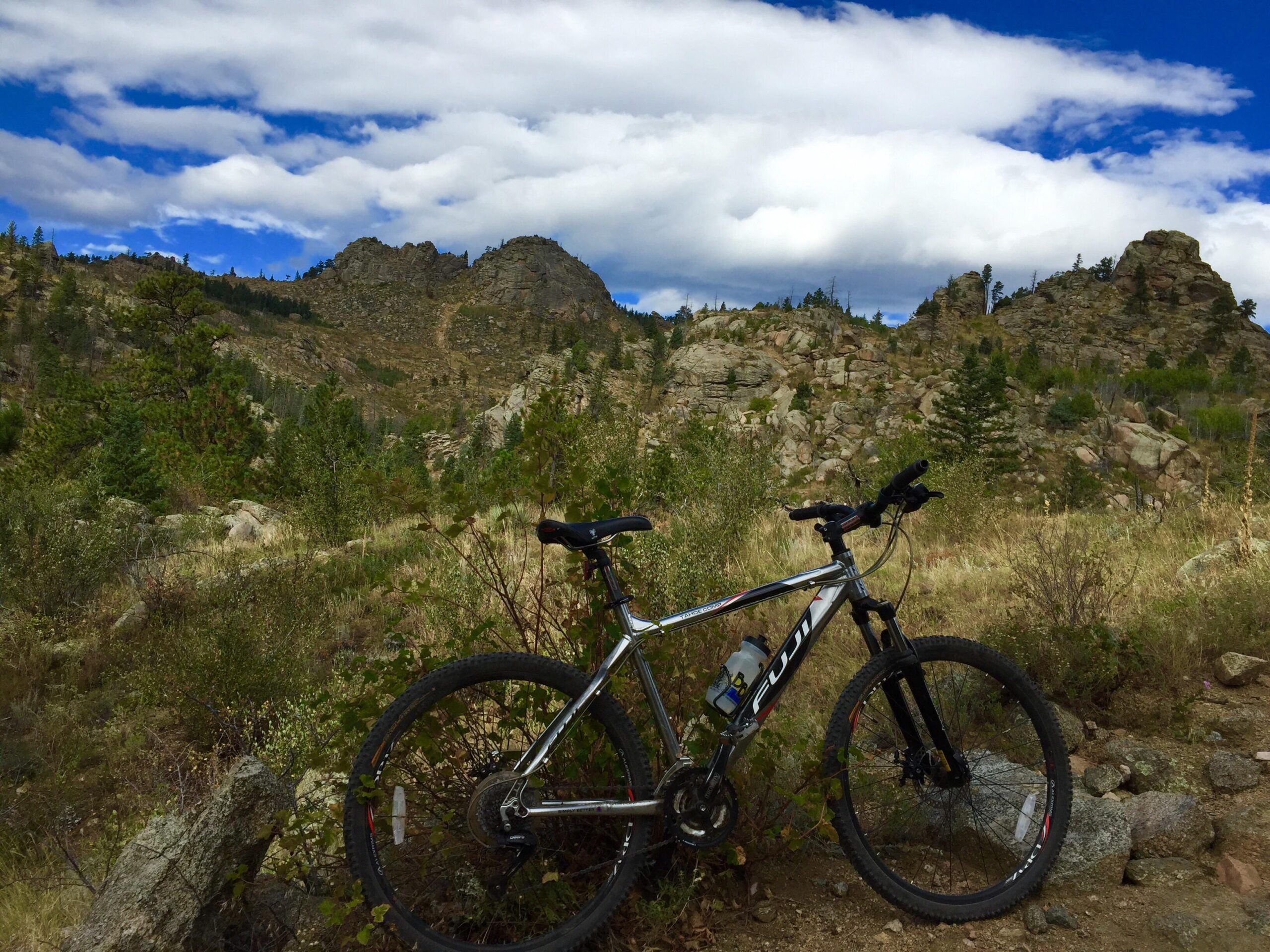 Mountain bike resting on a rocky trail, surrounded by lush greenery and towering rock formations under a blue sky with fluffy white clouds. Walker Ranch mountain bike trail.