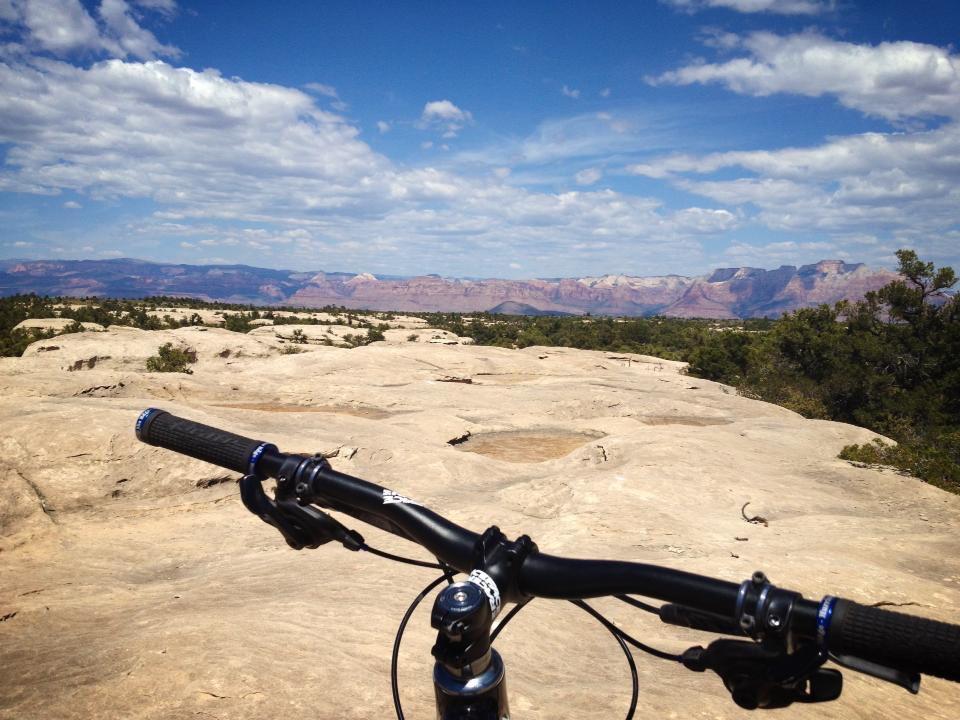 Mountain bike handlebars in the foreground with a vast rocky landscape and distant mountains under a bright blue sky with scattered clouds in the background. Gooseberry Mesa mountain bike trail.