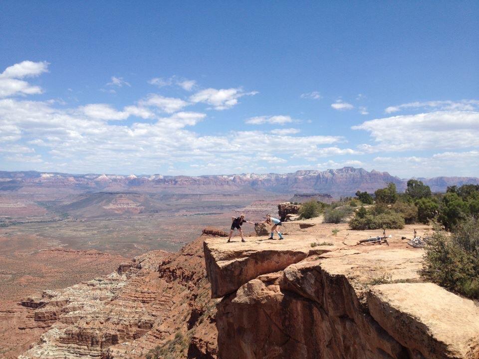 Two people engaging in an outdoor activity on a rocky ledge with a vast canyon and mountain landscape in the background. The sky is blue with scattered clouds, and there are bicycles nearby on the ground. Gooseberry Mesa mountain bike trail.
