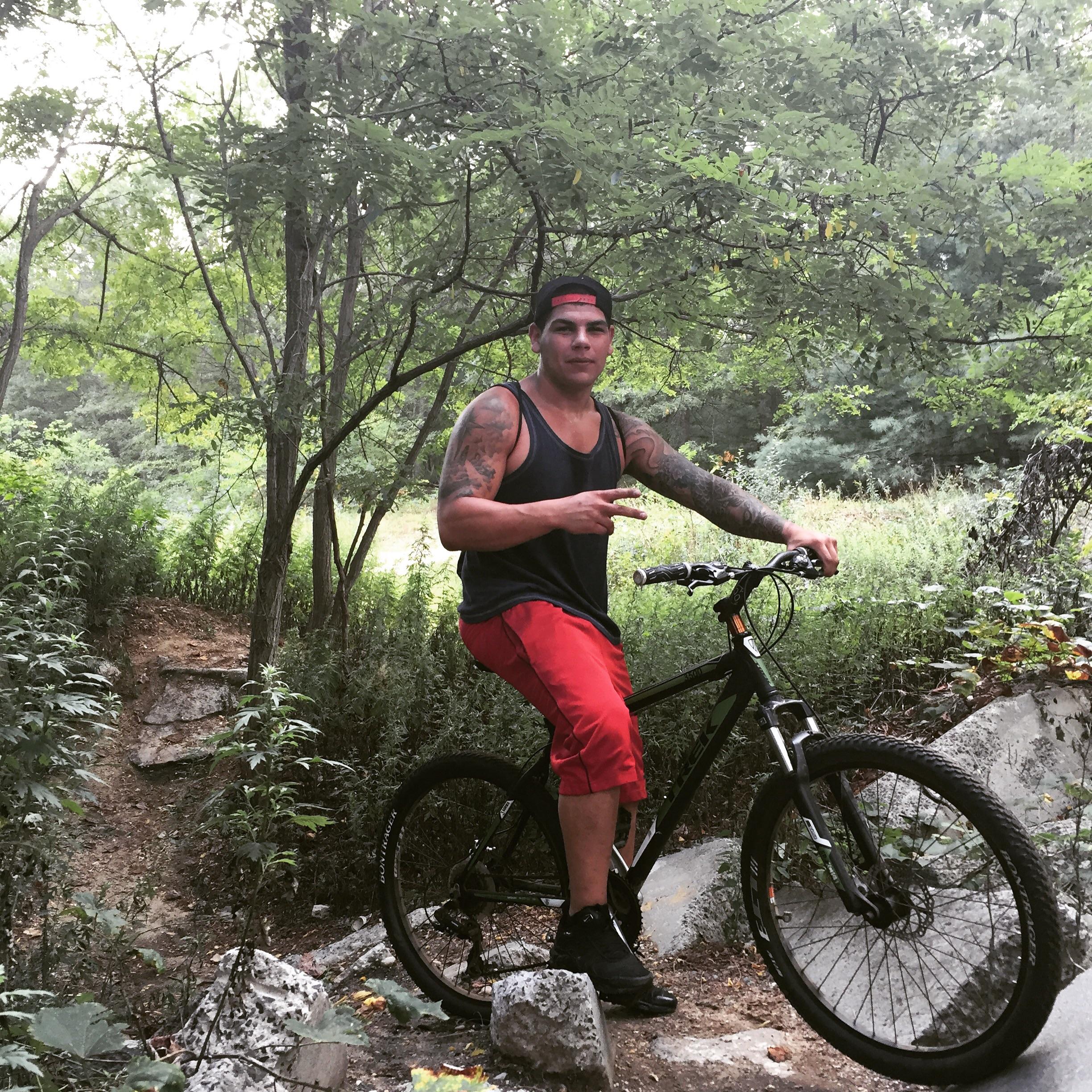 Trek 3500: A person with tattoos is sitting on a mountain bike in a wooded area, surrounded by greenery and rocks. They are wearing a black tank top, red shorts, and black shoes, and are making a peace sign with one hand while resting the other on the bike