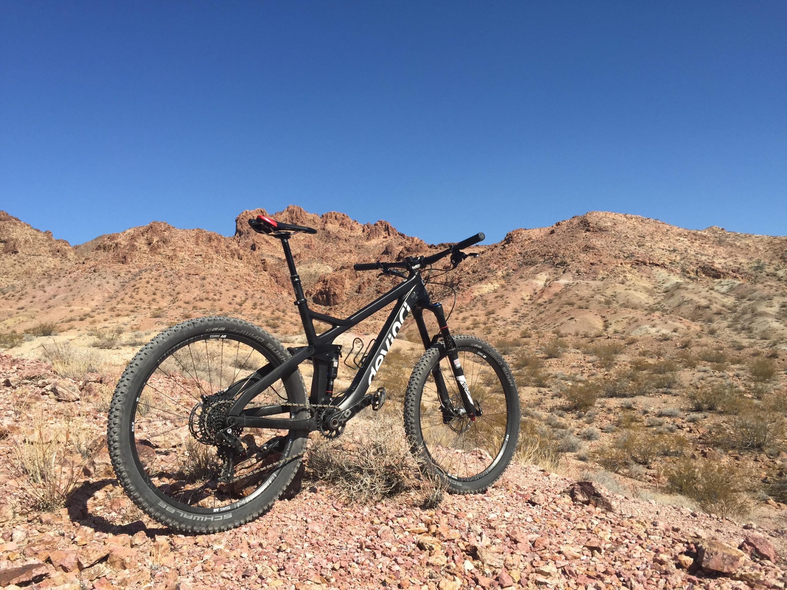 A black mountain bike leaning on rocky terrain with desert hills and a clear blue sky in the background. Bootleg Canyon mountain bike trail.