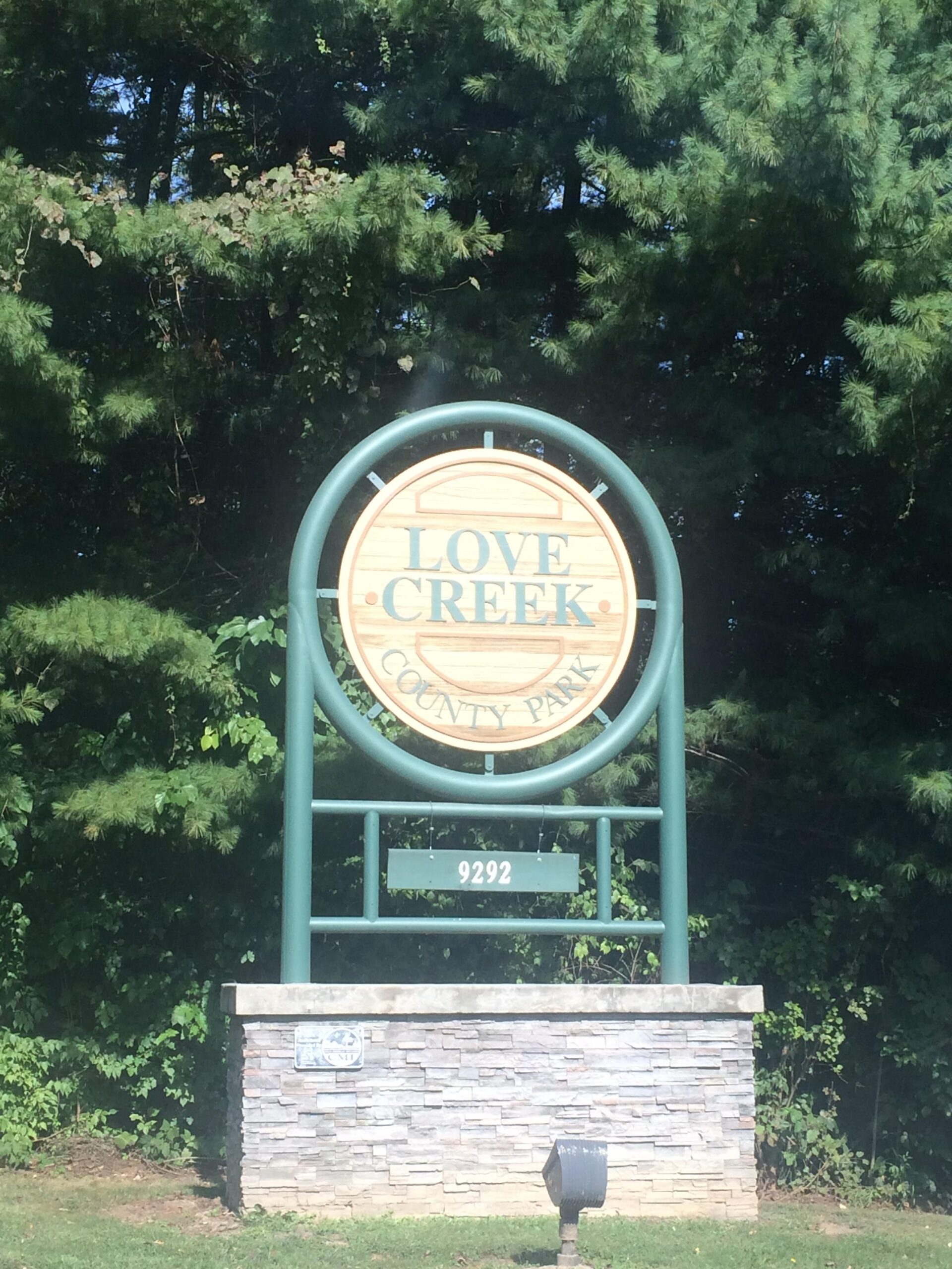 Sign for Love Creek County Park, featuring a wooden circular emblem with the text "LOVE CREEK" and "COUNTY PARK," surrounded by greenery and trees. The base of the sign includes a stone wall and a plaque with the address number "9292." Love Creek mountain bike trail.