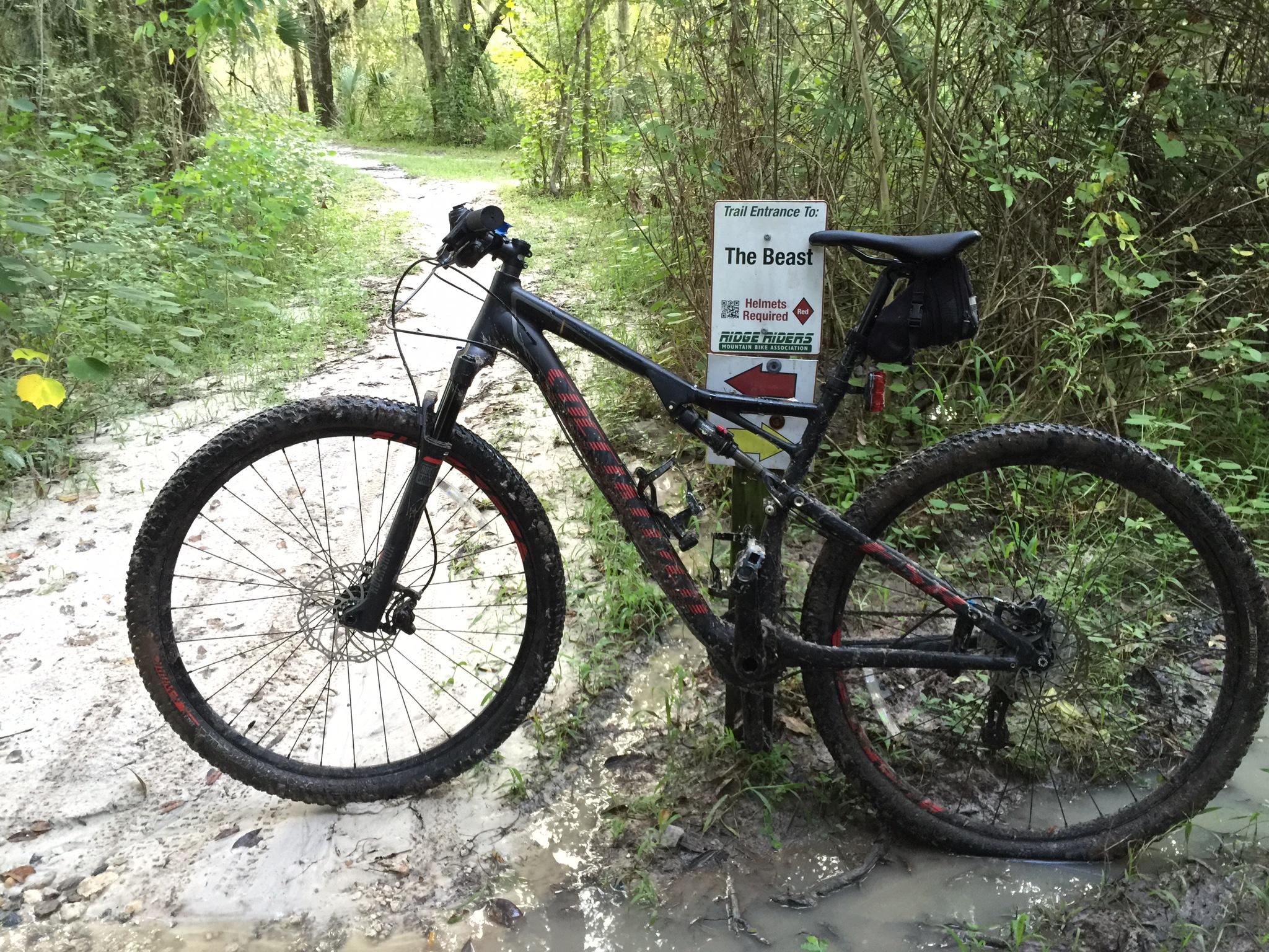 Specialized Epic: A mountain bike resting on a sandy trail with muddy tires, next to a sign that reads "Trail Entrance To: The Beast," indicating the start of a biking trail. Lush green foliage surrounds the path, suggesting a natural outdoor environment.