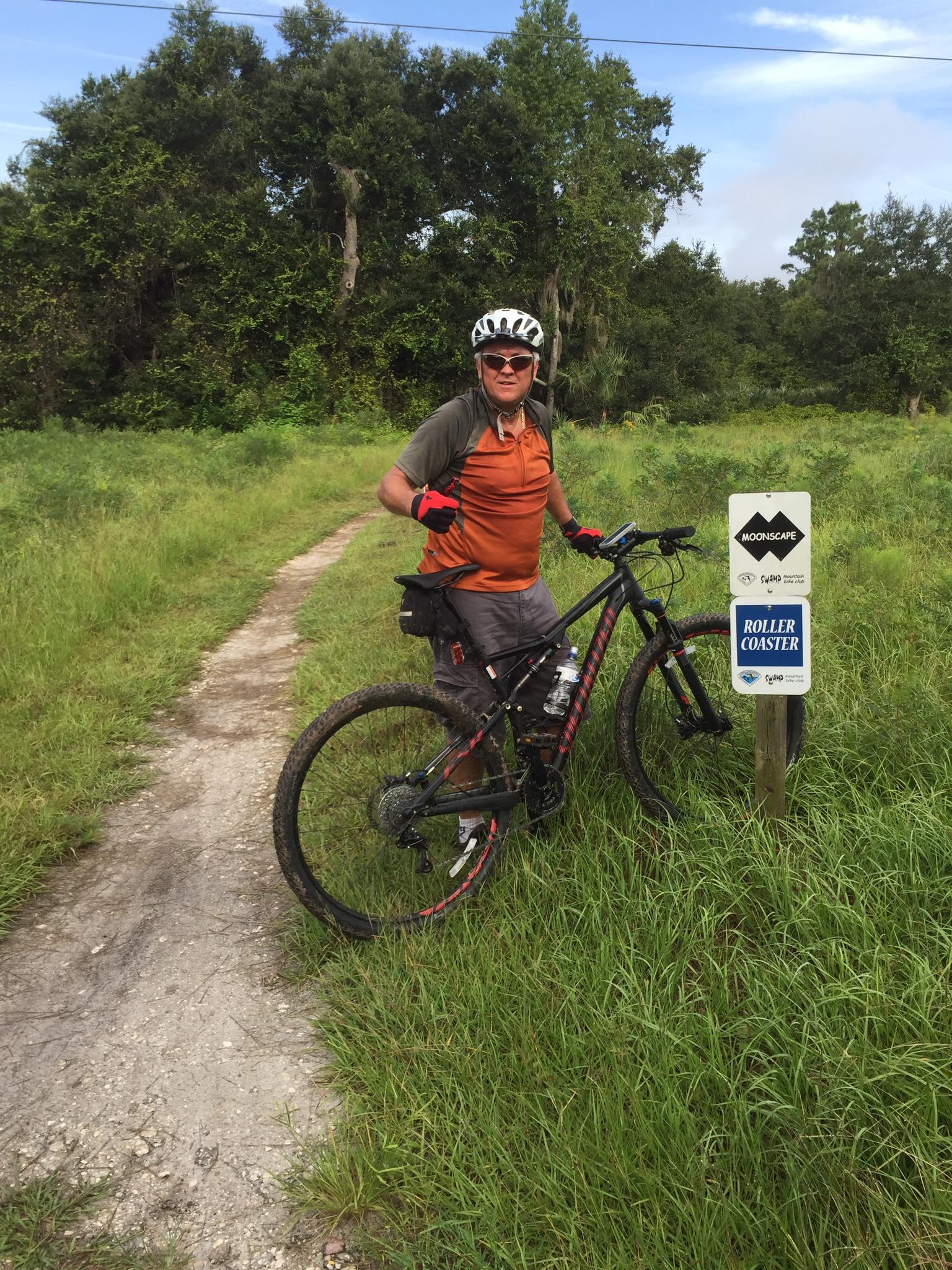 Specialized Epic: A person wearing a helmet and orange shirt stands next to a mountain bike on a dirt path surrounded by tall grass and trees. They are giving a thumbs-up gesture, with a trail sign that reads "Moonscape" and mentions "Roller Coaster" in the background.