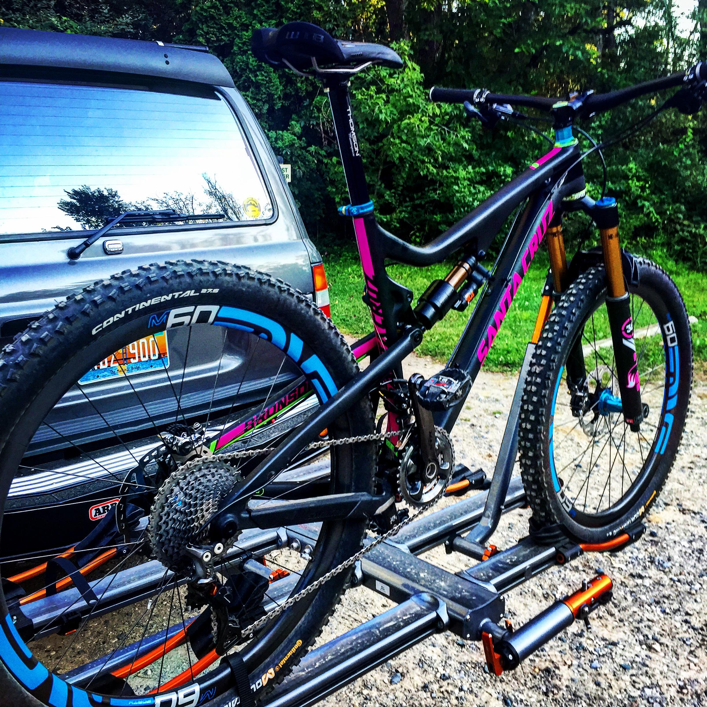 Santa Cruz Bronson: A colorful mountain bike is mounted on a bike rack attached to a vehicle, with lush greenery in the background. The bike features a black frame with vibrant pink and blue accents, chunky tires, and a visible drivetrain. The rear of the vehicle is partially visible, showcasing its window and bumper.