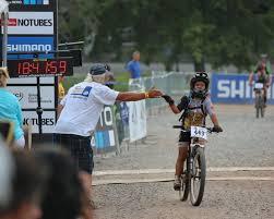 A young cyclist crossing the finish line of a race, celebrating with a supportive adult who is giving them a high five. The scene takes place on a gravel path with a race timer visible in the background. Both the cyclist and the adult appear joyful and encouraging.