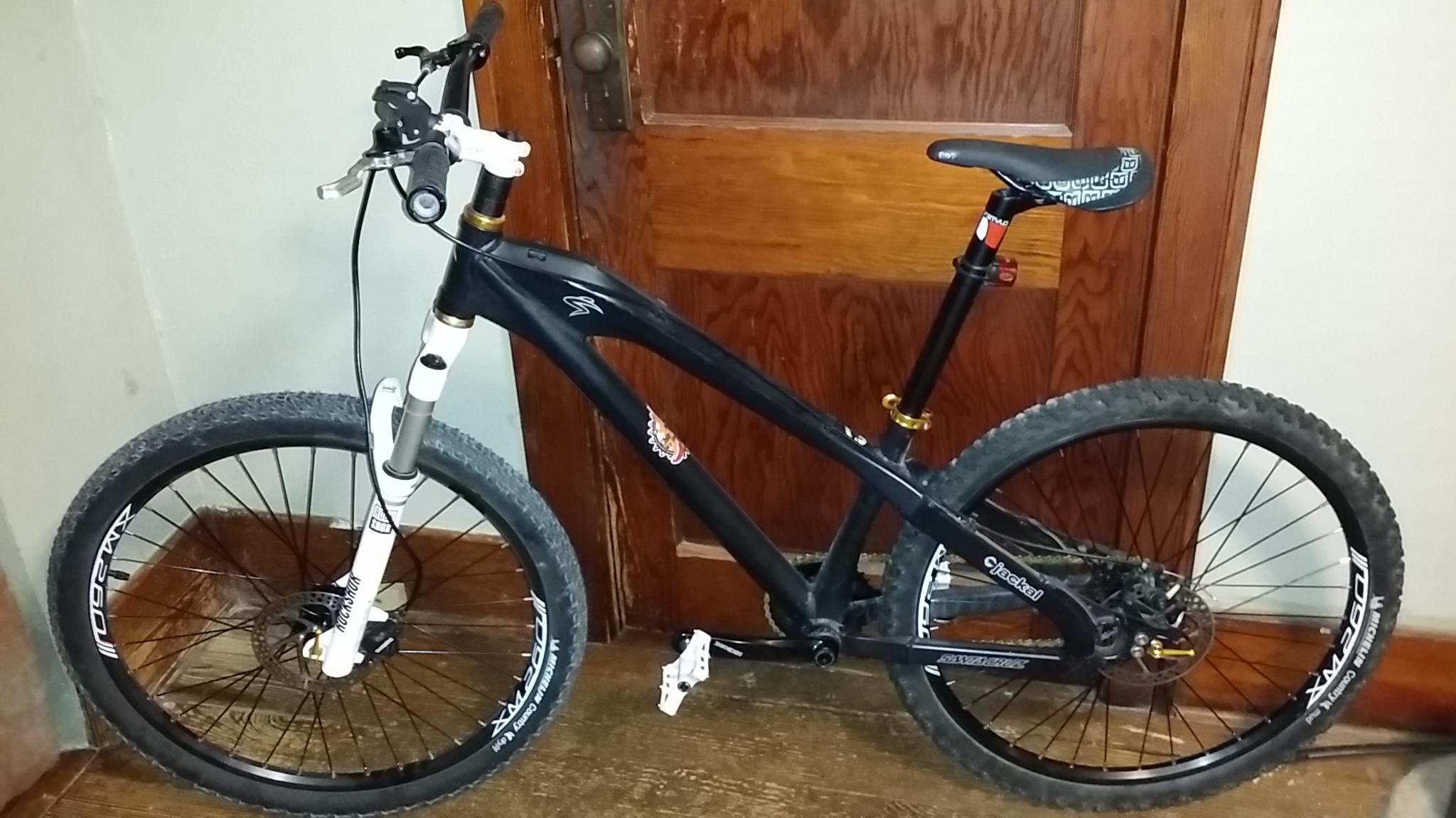 Santa Cruz Jackal: A black mountain bike leaning against a wooden door, featuring a white front fork and thick, textured tires. The bike has a black seat with a patterned design and is equipped with disc brakes. The background shows a wooden floor and an interior wall.